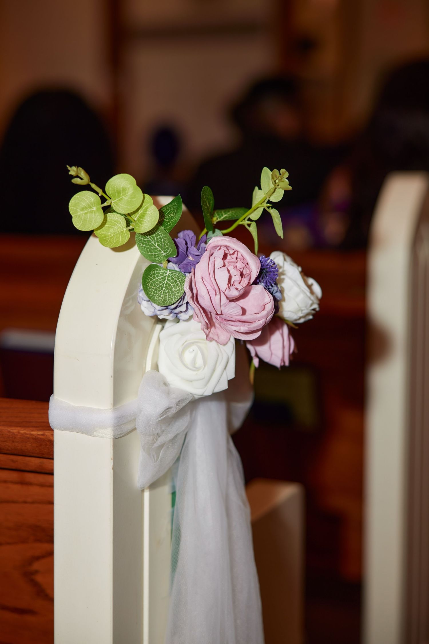 Elegant wedding pew decoration with pink, white, and purple flowers, green accents, and white ribbon in a church.
