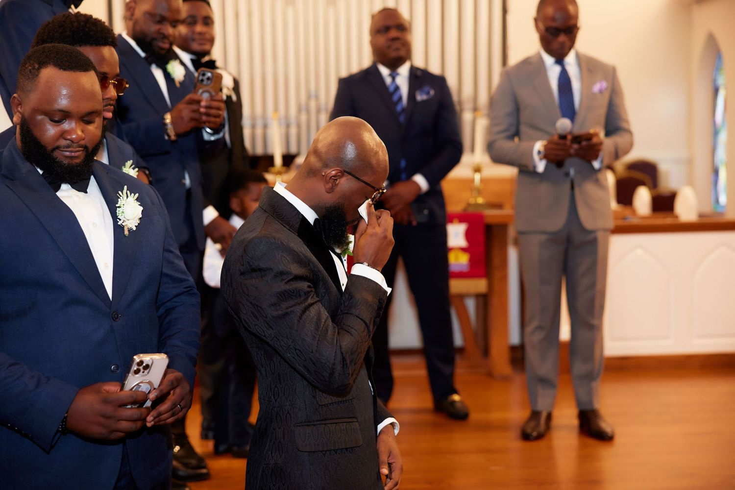 Emotional groom wiping tears at wedding ceremony surrounded by groomsmen in navy suits inside a church.