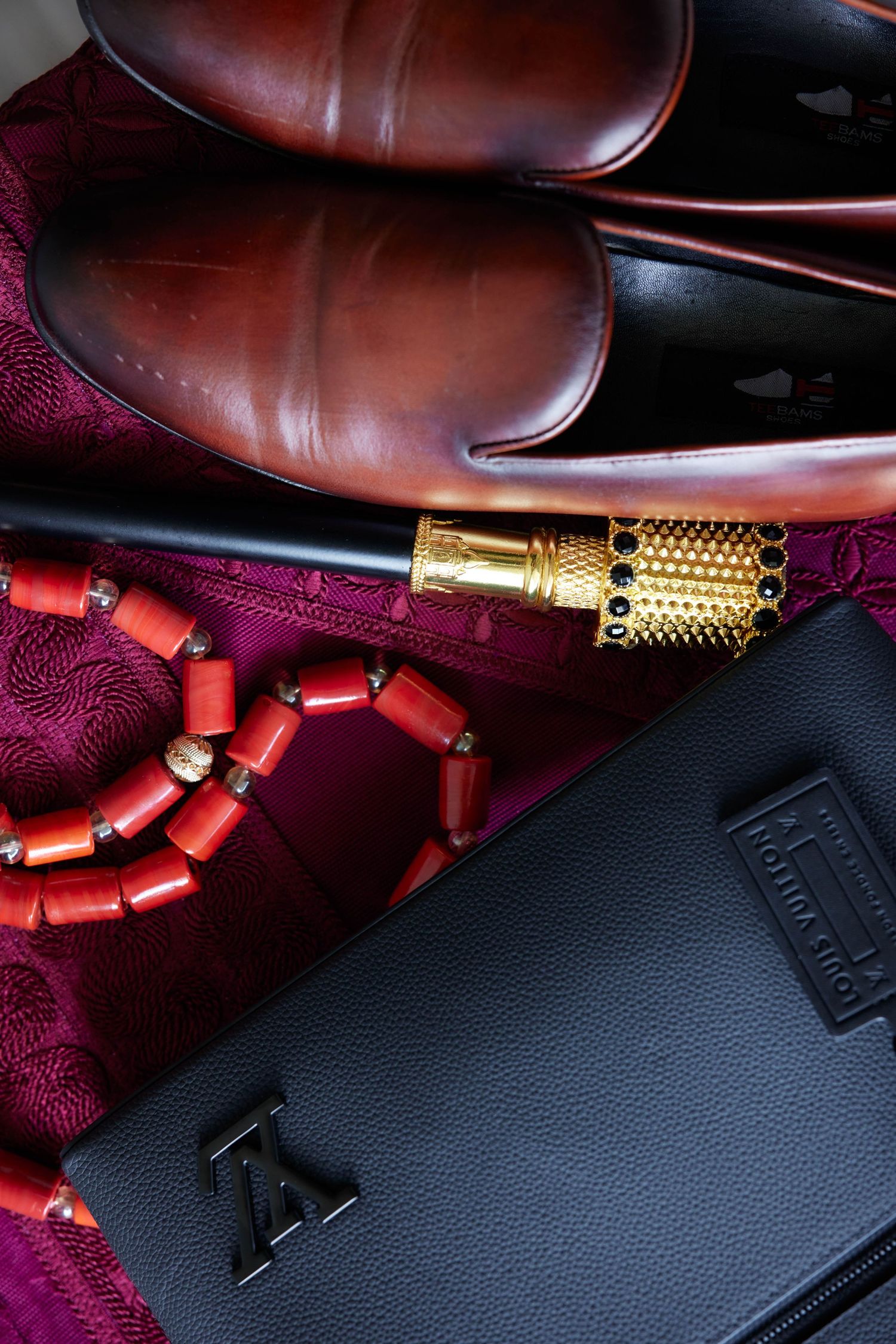 Luxury flat lay with brown leather loafers, red coral beads, gold lipstick, and black Louis Vuitton wallet on purple fabric.