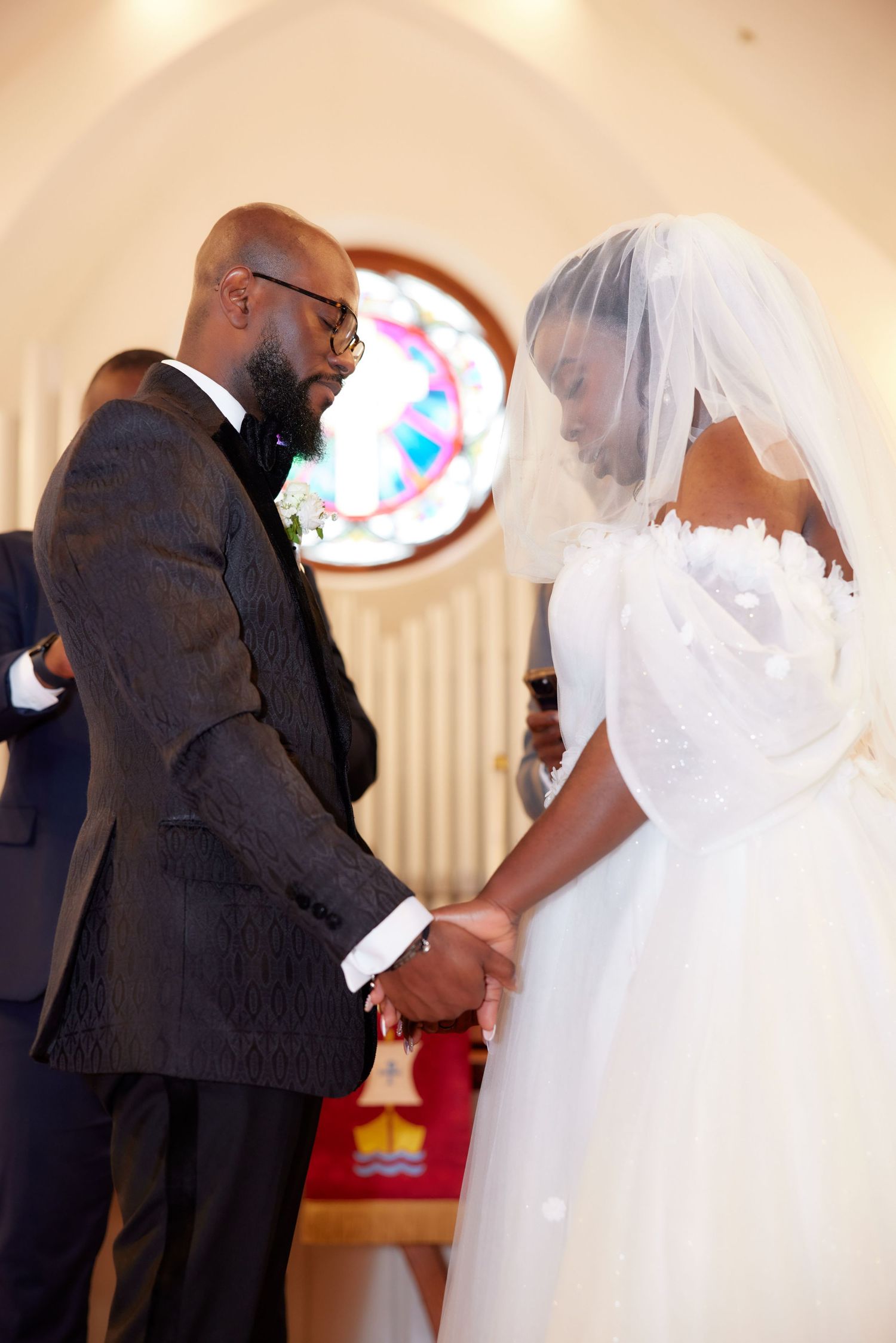 Bride and groom holding hands at church altar during wedding ceremony, stained glass window in background.