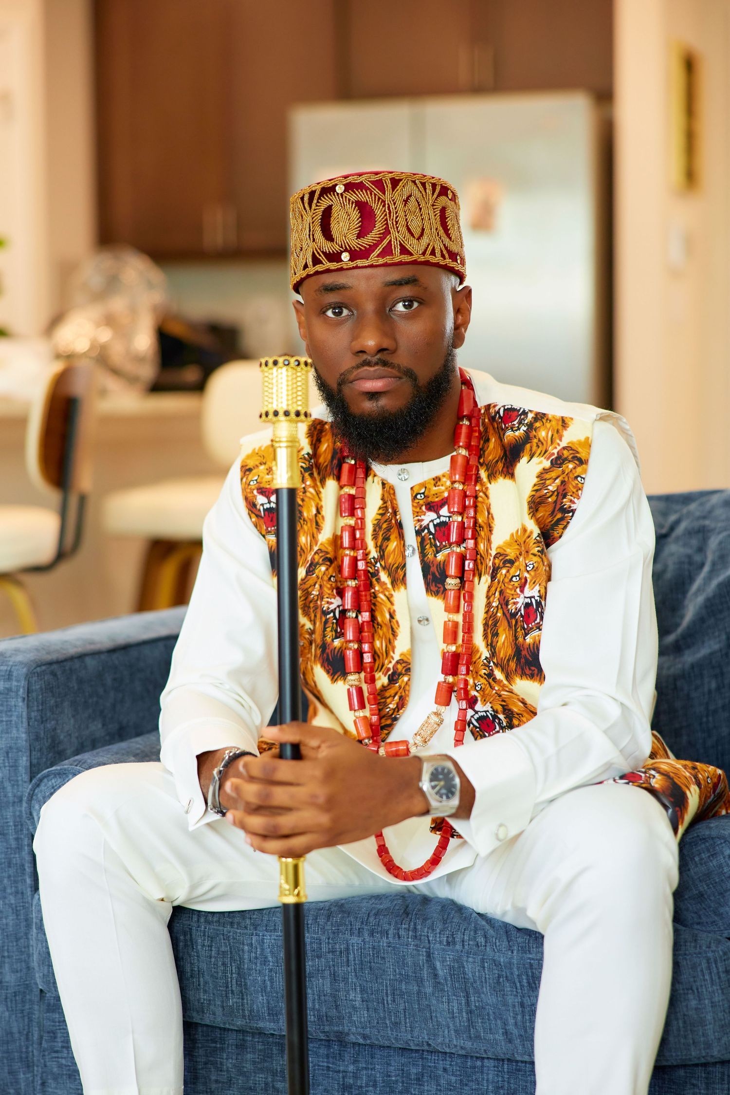 Man in traditional African attire with red beaded necklace, ornate cap, and golden staff seated on a blue sofa.