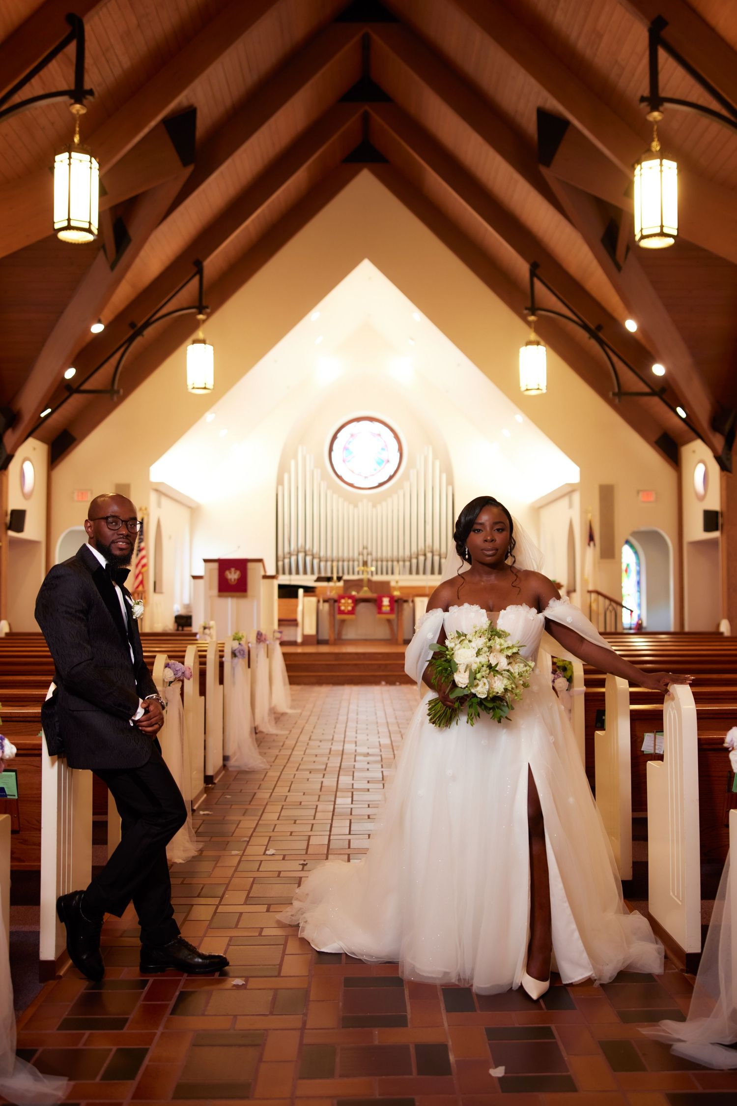 Bride in white off-shoulder gown and groom in tuxedo pose in elegant church aisle.