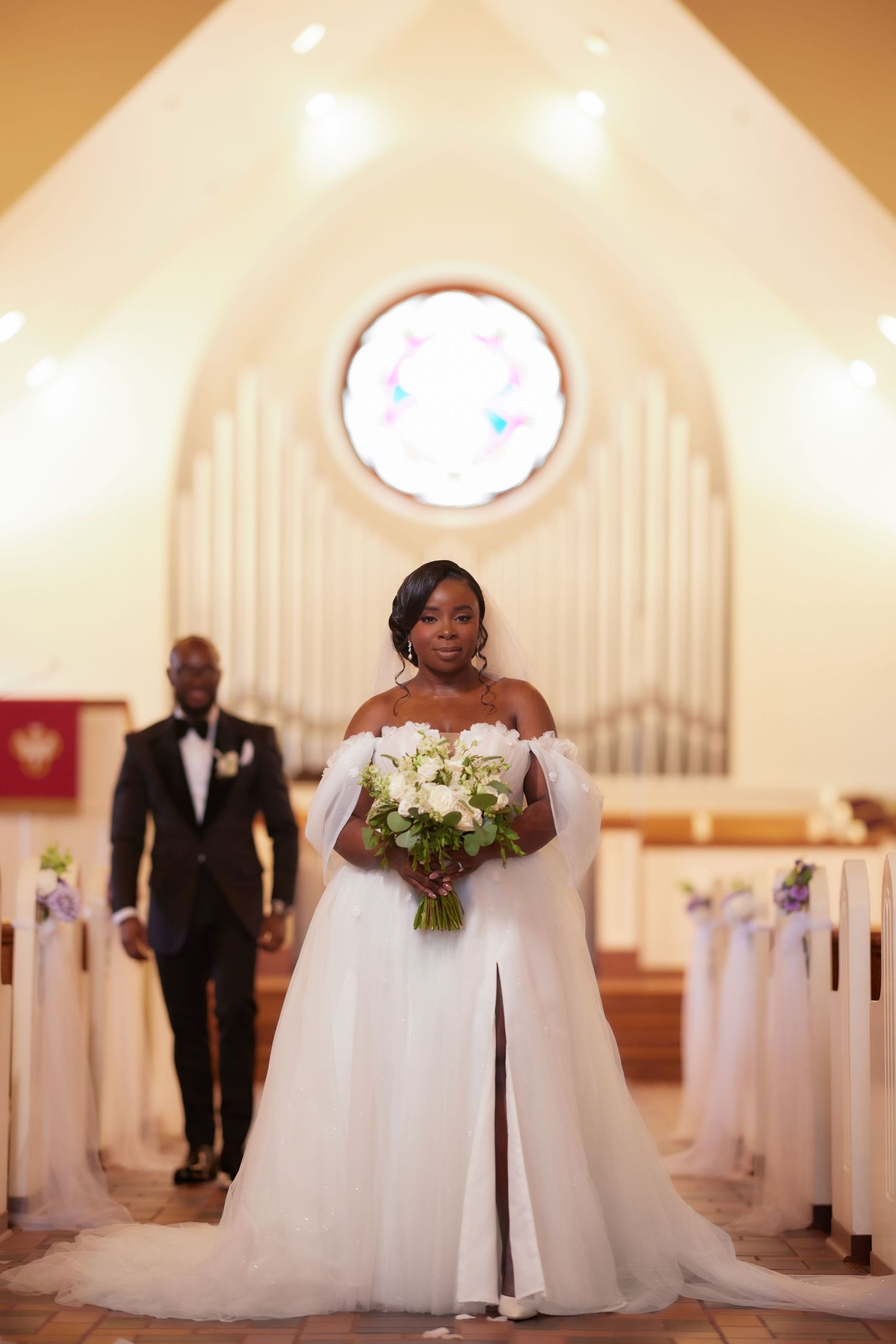 Bride in white off-shoulder gown holding bouquet walks down church aisle toward groom.