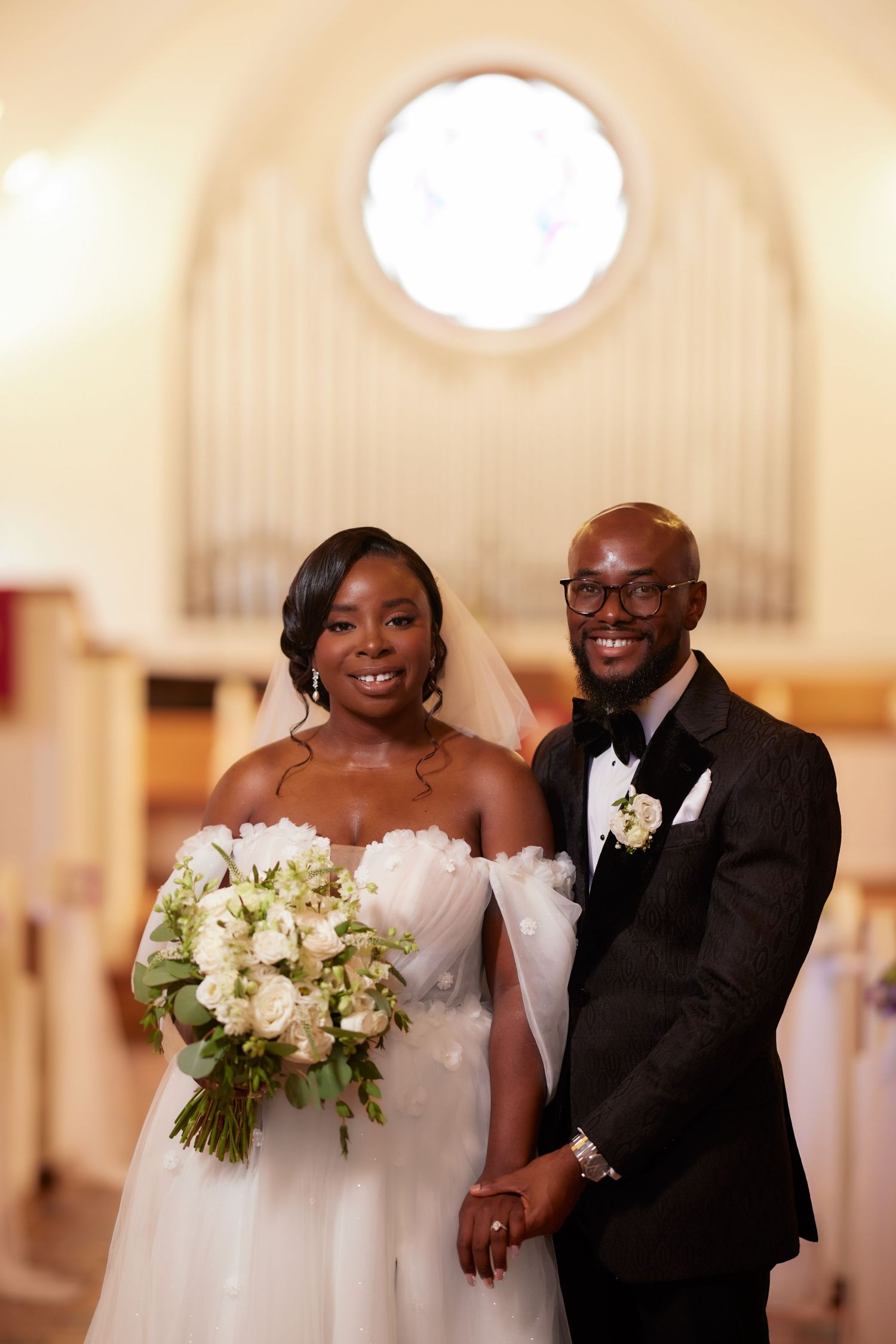 Smiling newlywed couple posing in church aisle, bride in white gown with bouquet, groom in black tuxedo.