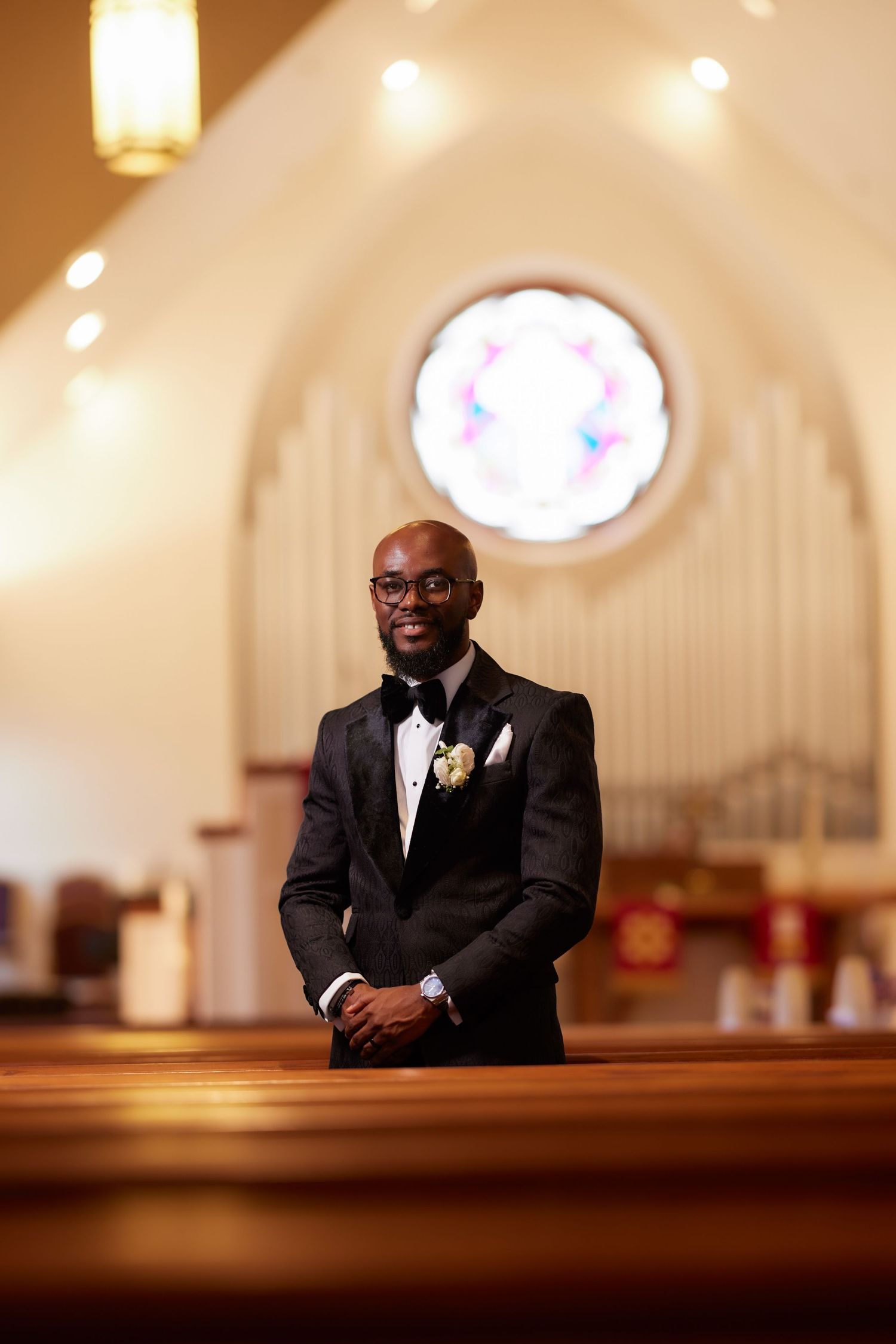 Smiling groom in black tuxedo with boutonniere stands in church aisle before wedding ceremony.