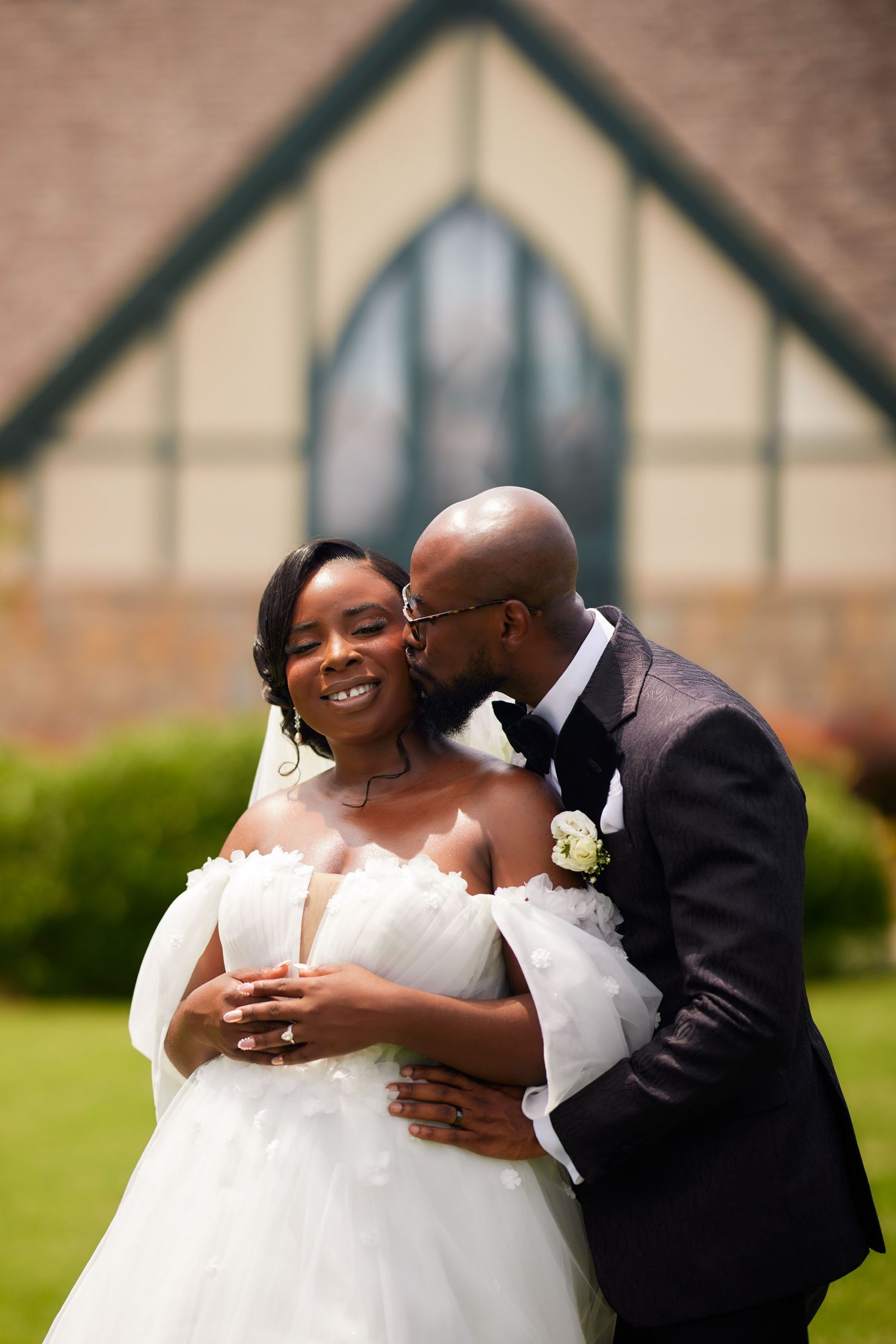 Groom kisses bride on cheek outside church, bride smiles in off-shoulder white gown, groom in dark suit with boutonniere.