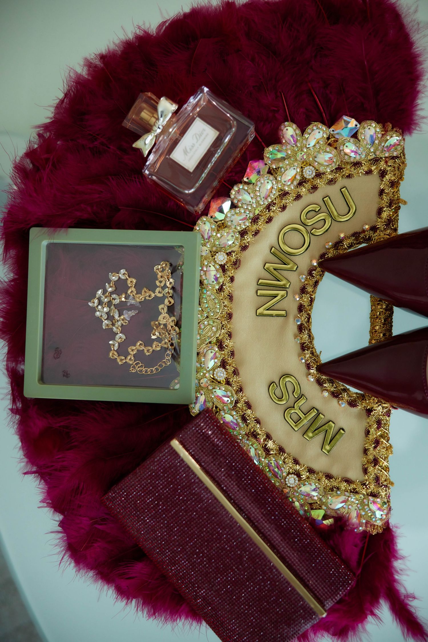 Bridal flat lay with Mrs. Brown crown, gold jewelry box, burgundy clutch, and perfume on pink fur.