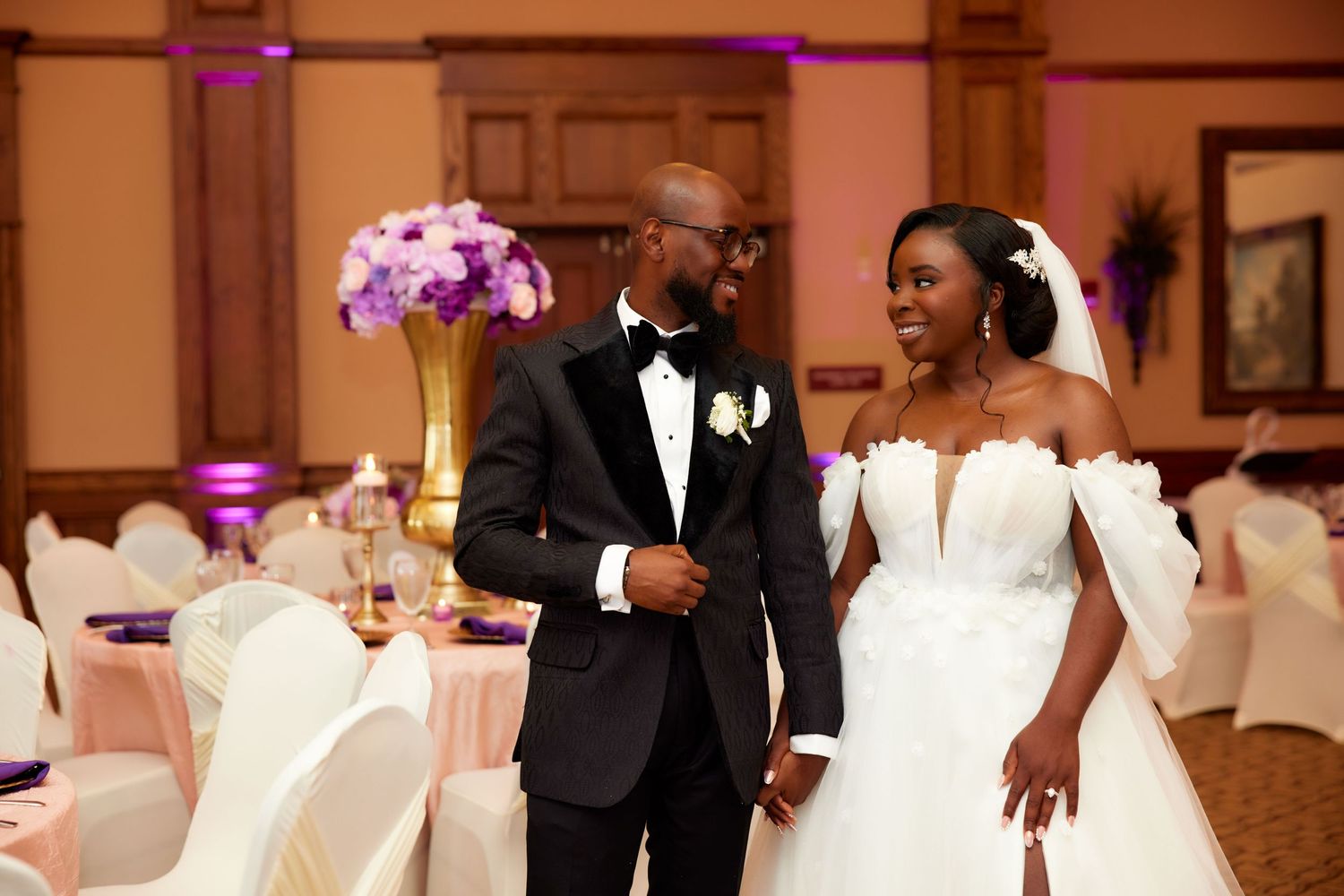 Elegant bride and groom in wedding attire smile at each other in a beautifully decorated ballroom reception venue.