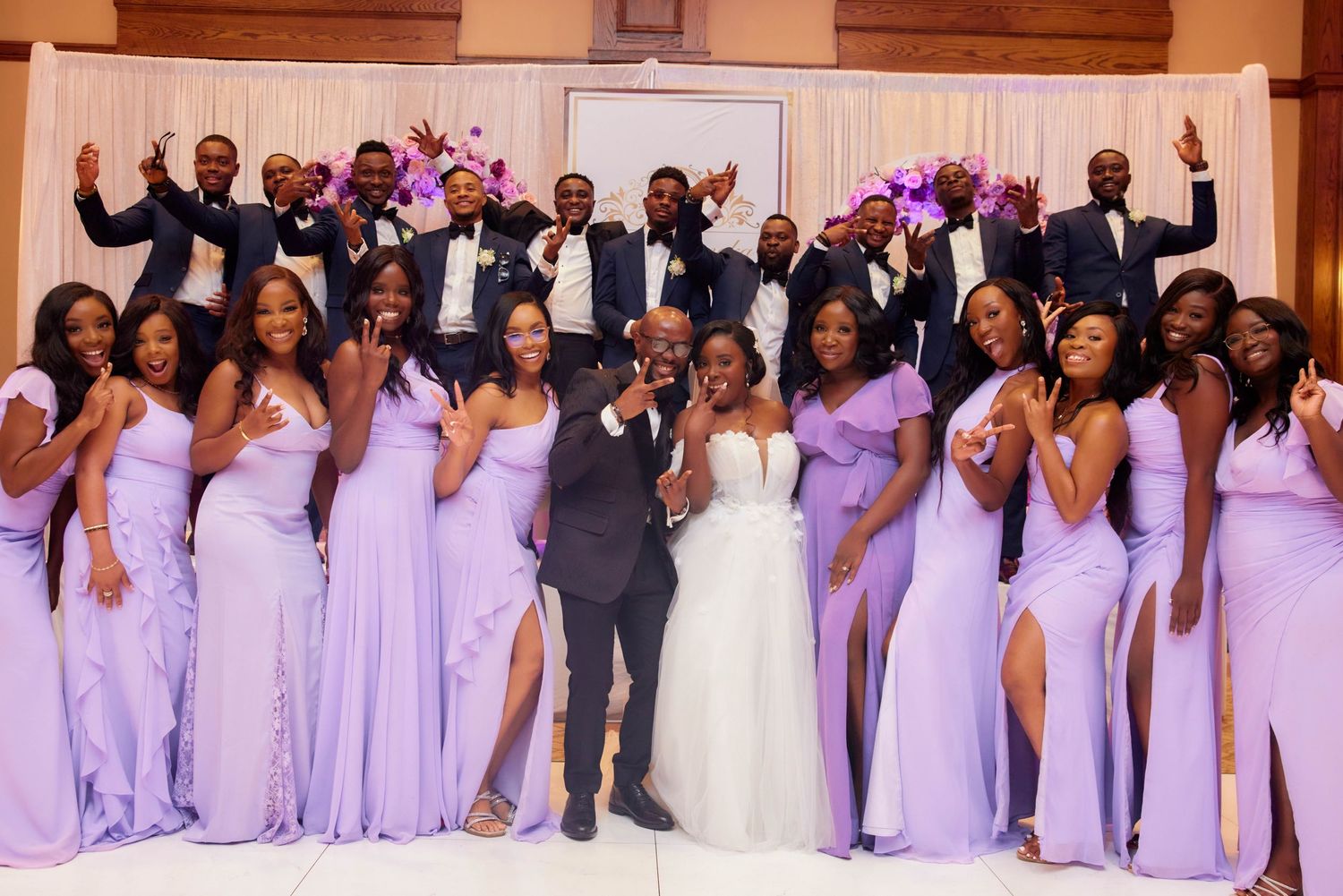 Wedding party poses together, bride and groom surrounded by bridesmaids in lavender gowns and groomsmen in navy suits.