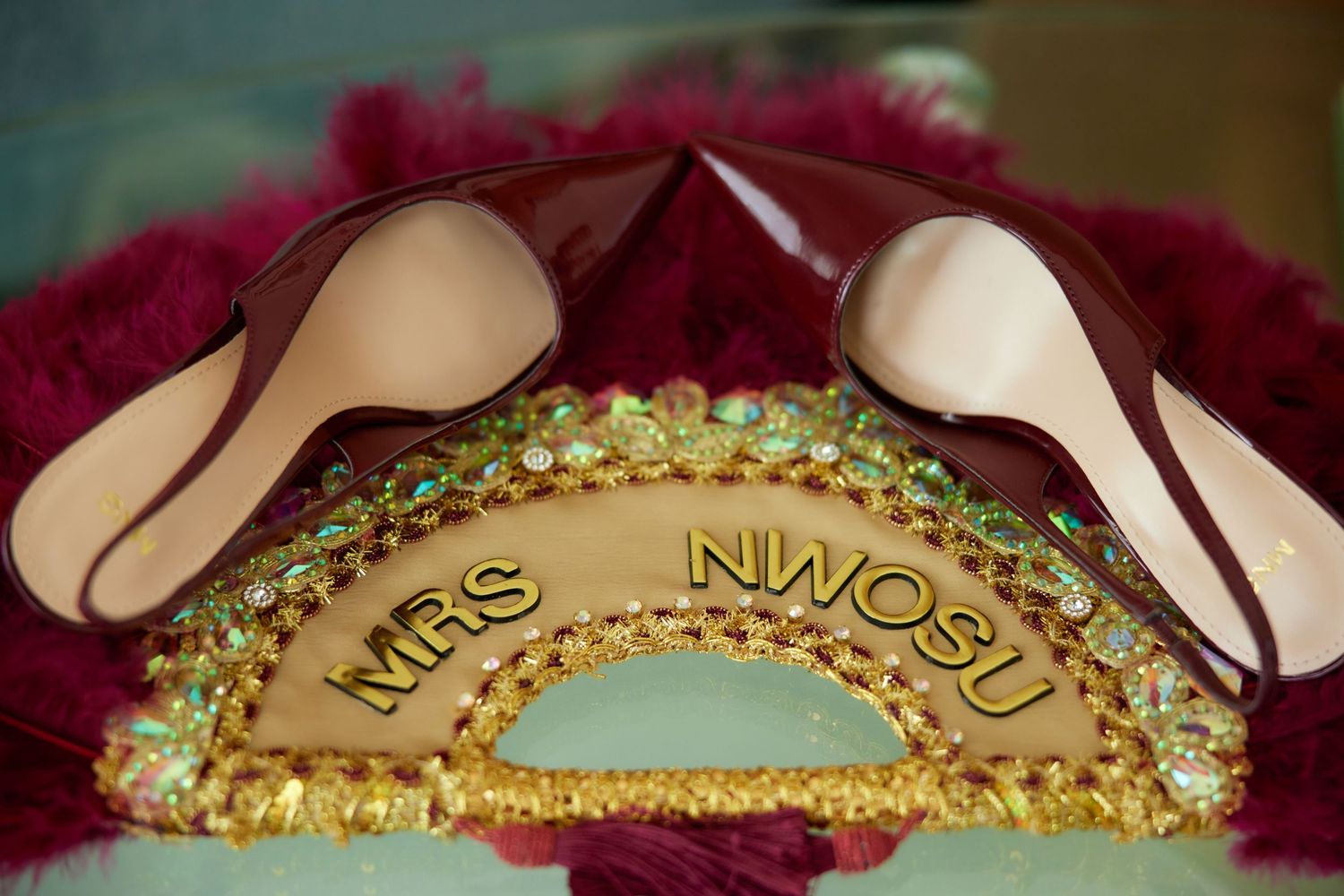 Gold jeweled Mrs. Nwosu wedding crown with pink satin heels displayed on burgundy feathered backdrop.
