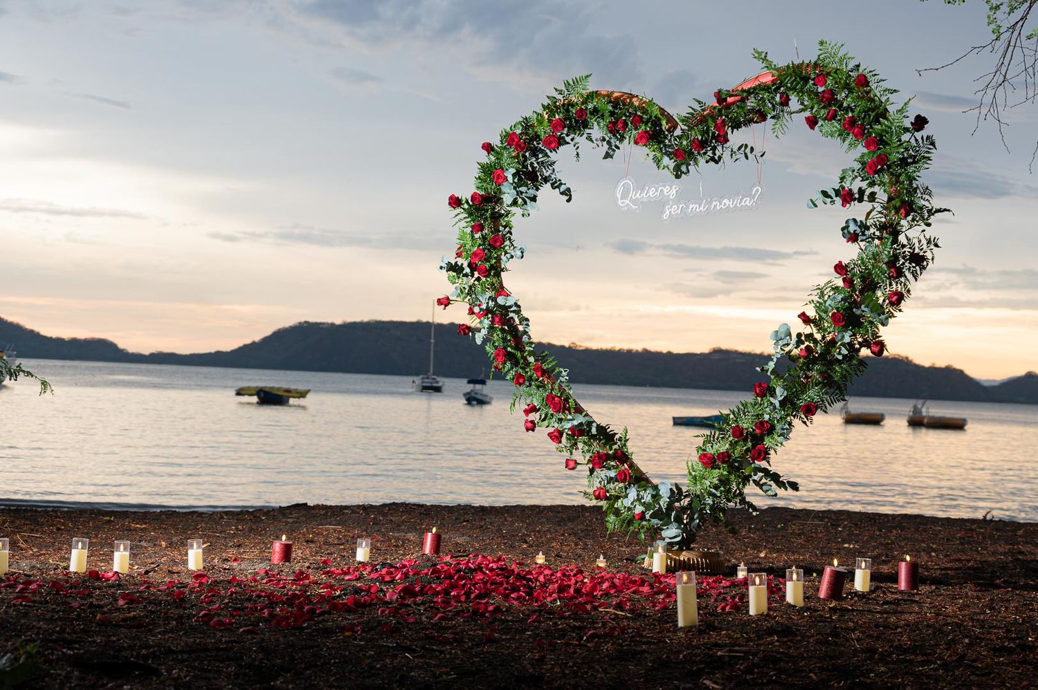 Heart-shaped floral arch with red roses on a beach at sunset, with candles and calm water in the background.