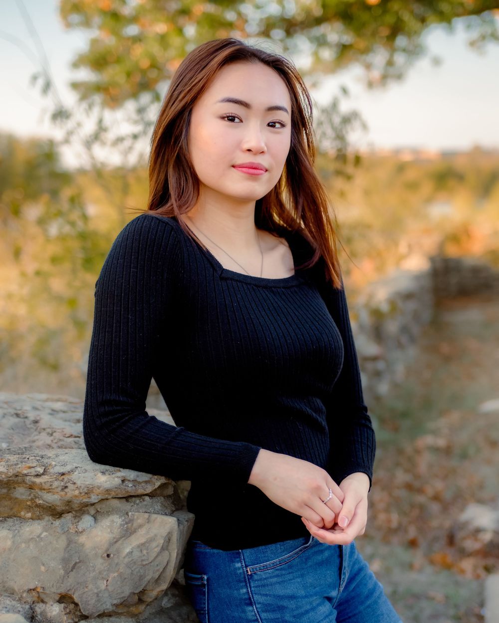 Young woman in black sweater and jeans posing outdoors against a stone wall with autumn foliage background.