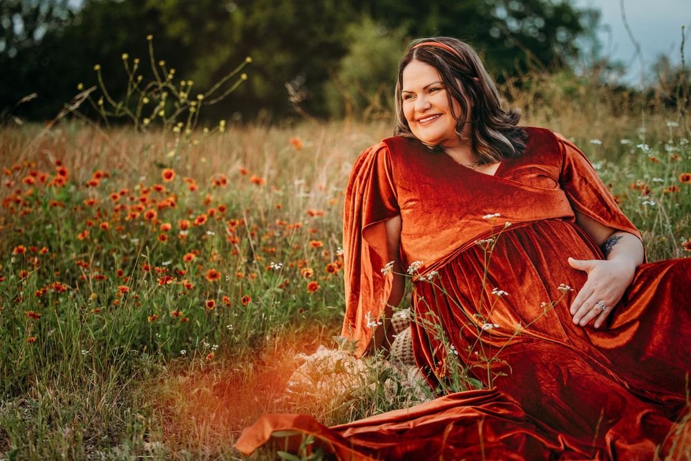 Pregnant woman in red velvet gown sitting in wildflower field with poppies during maternity photo session.