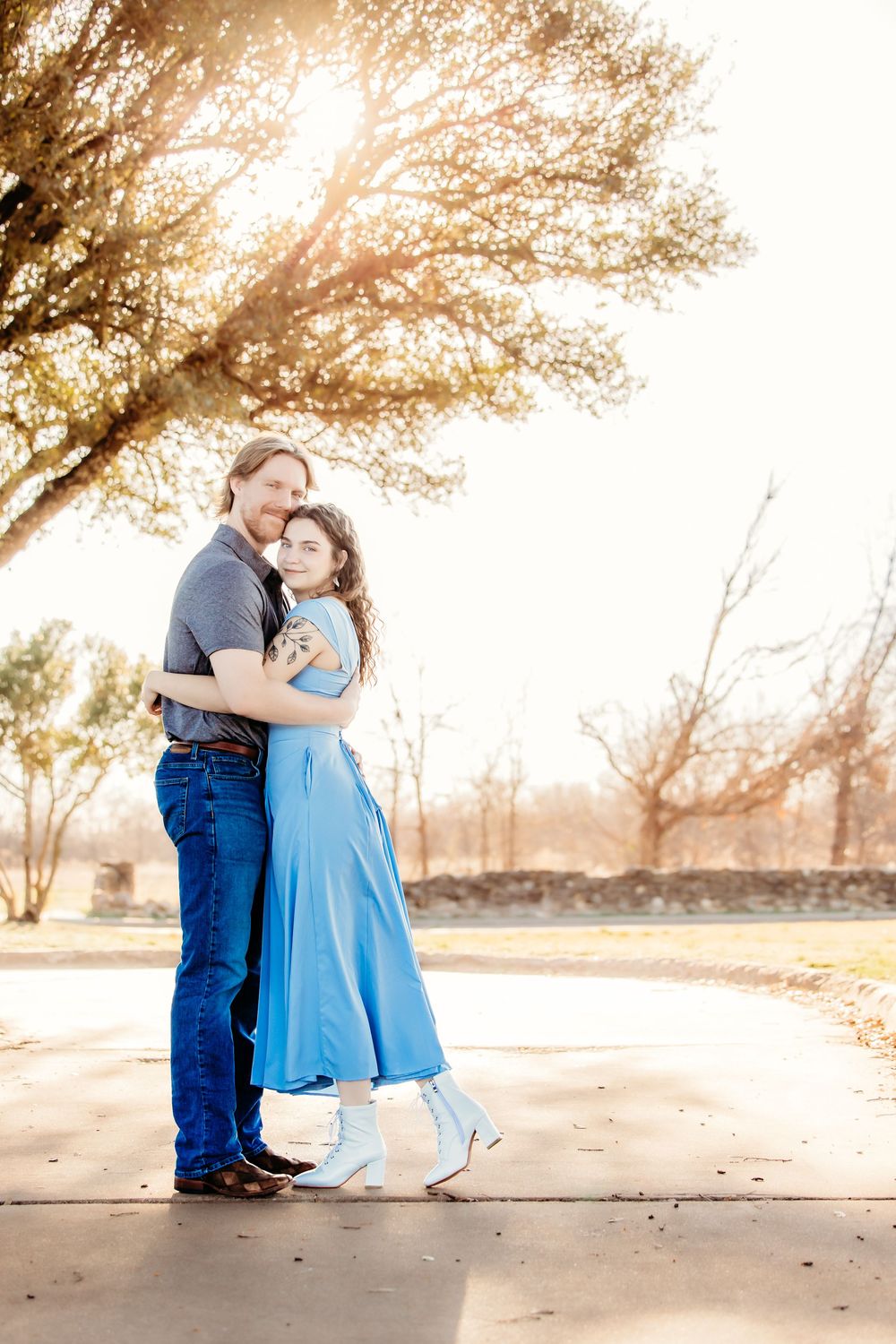 Couple embracing outdoors in golden sunlight, woman in blue dress and white boots, man in jeans, near a tree in a park.