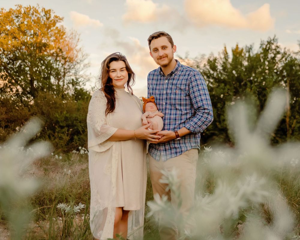 A couple stands outdoors at golden hour, smiling and holding a baby dressed in orange, surrounded by lush greenery.