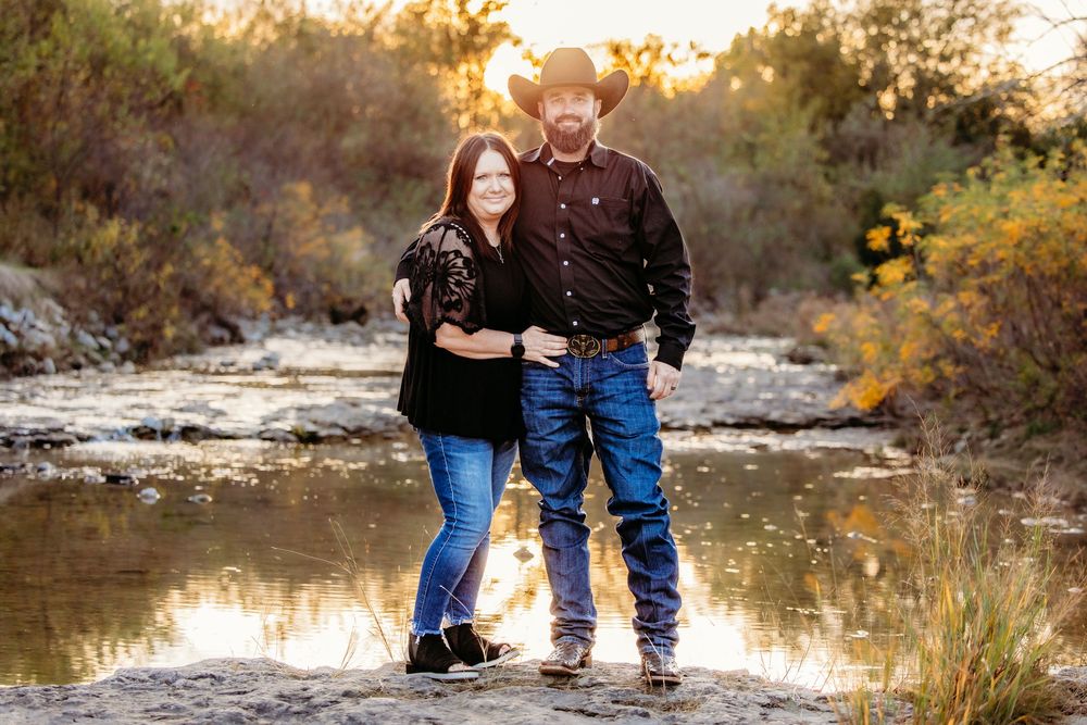 Couple posing together near a creek at sunset, wearing black shirts and jeans in an outdoor fall setting.