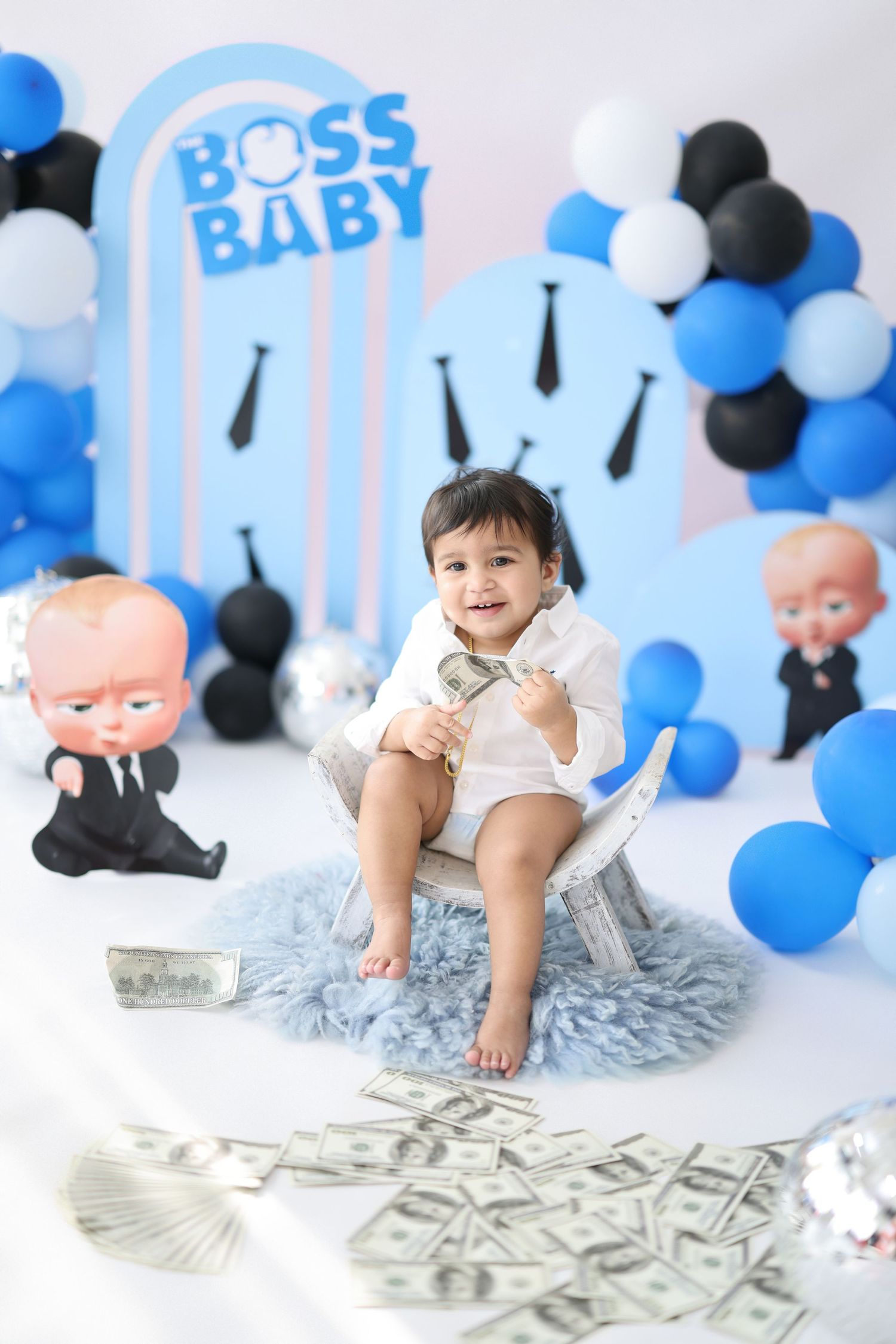 Baby boy sits on chair with Boss Baby themed backdrop, blue balloons, and dollar bills for first birthday cake smash.