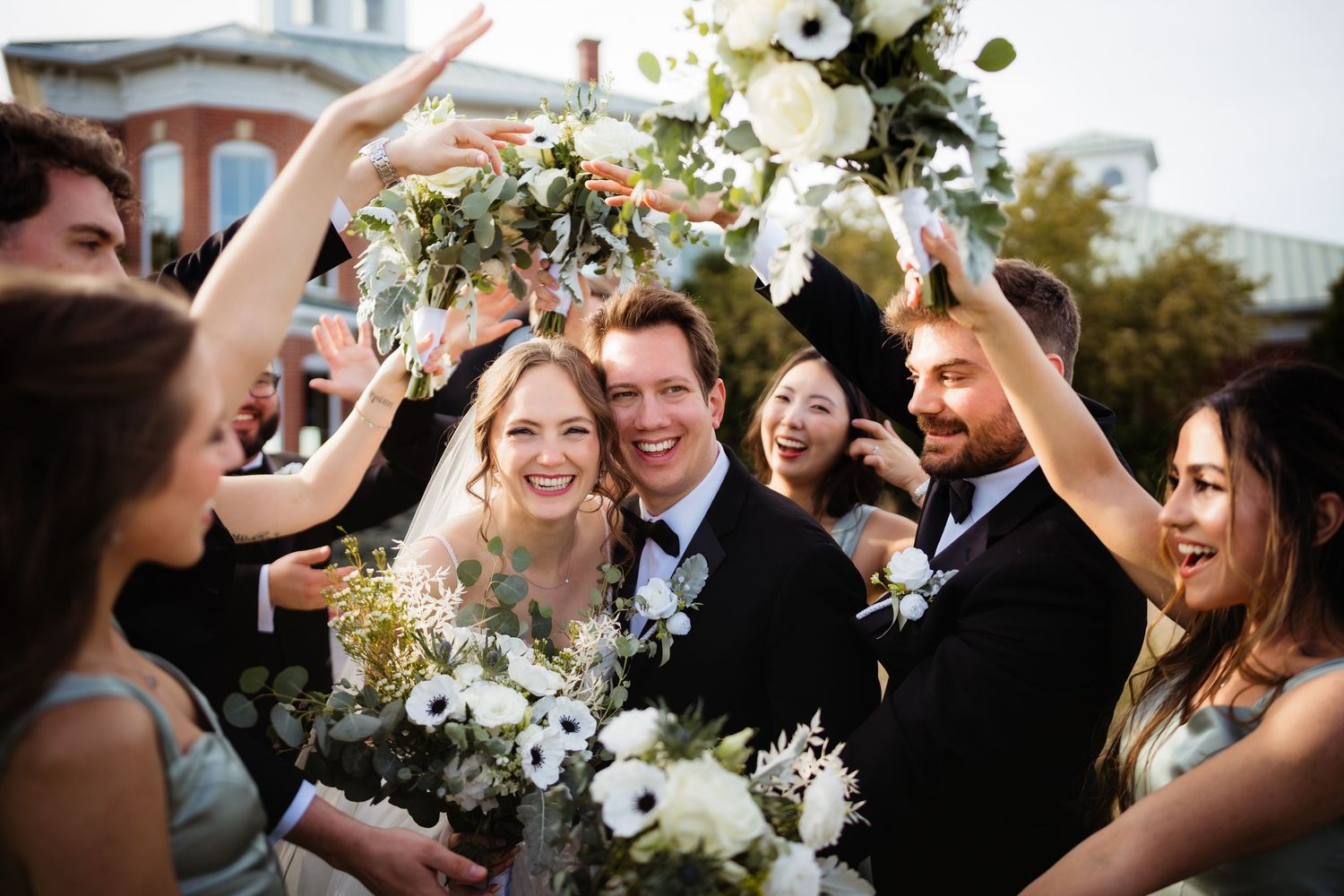 Joyful bride and groom surrounded by wedding party celebrating outdoors with white floral bouquets and greenery.