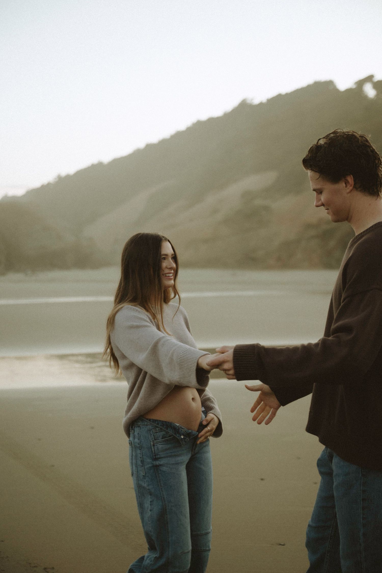 Couple holding hands and smiling on a misty beach with mountains in the background.