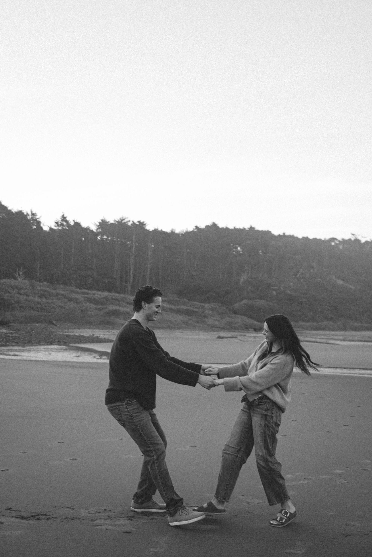 Black and white photo of a couple holding hands and dancing playfully on a beach with trees in the background.