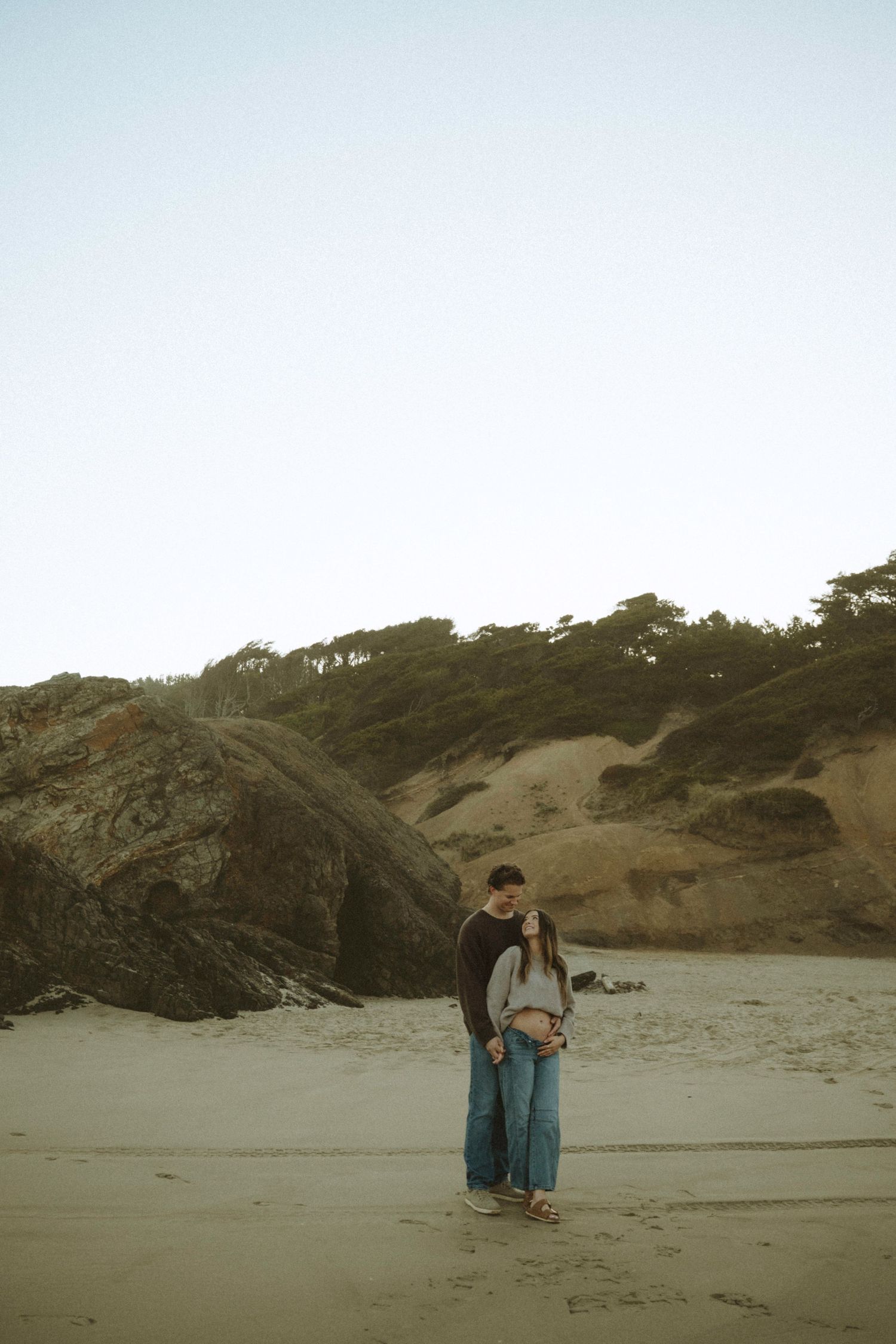 Couple embracing romantically on a sandy beach with rocky cliffs and trees in the background.