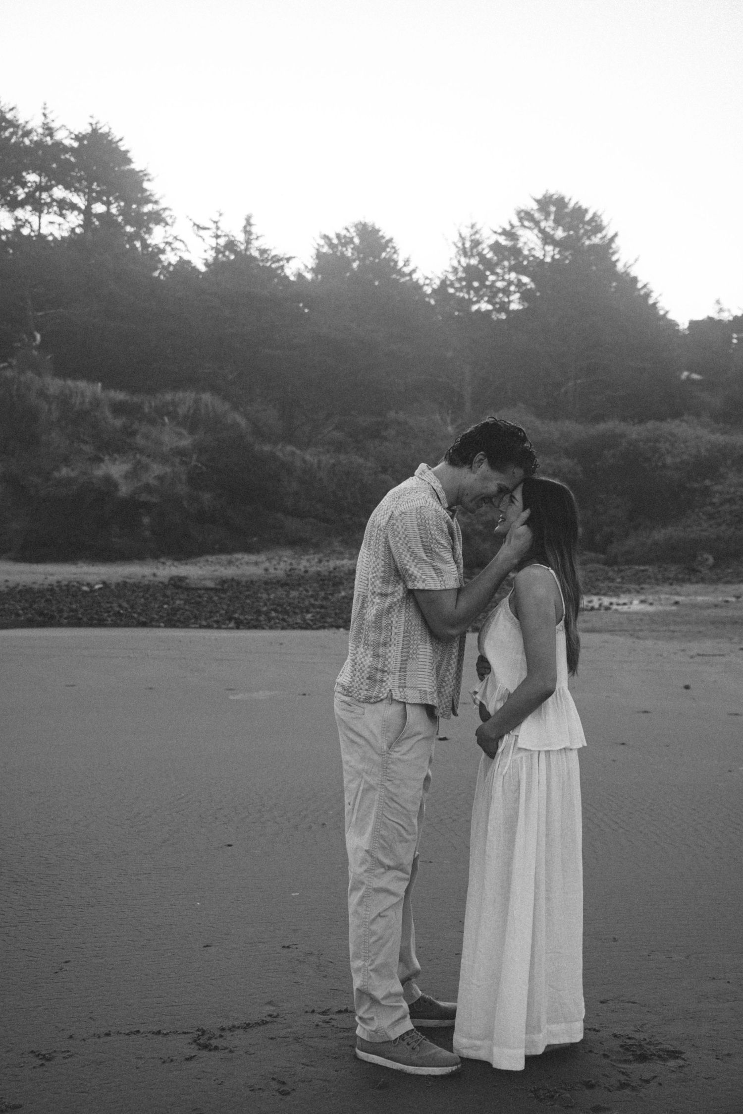 Black and white photo of a romantic couple kissing on a beach with trees in the background.