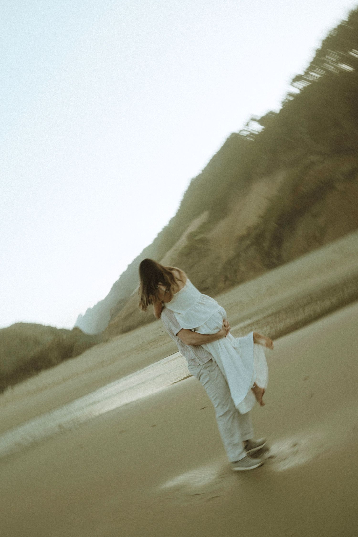 A woman in white dress and pants dances gracefully on a sandy beach with coastal cliffs in the background.