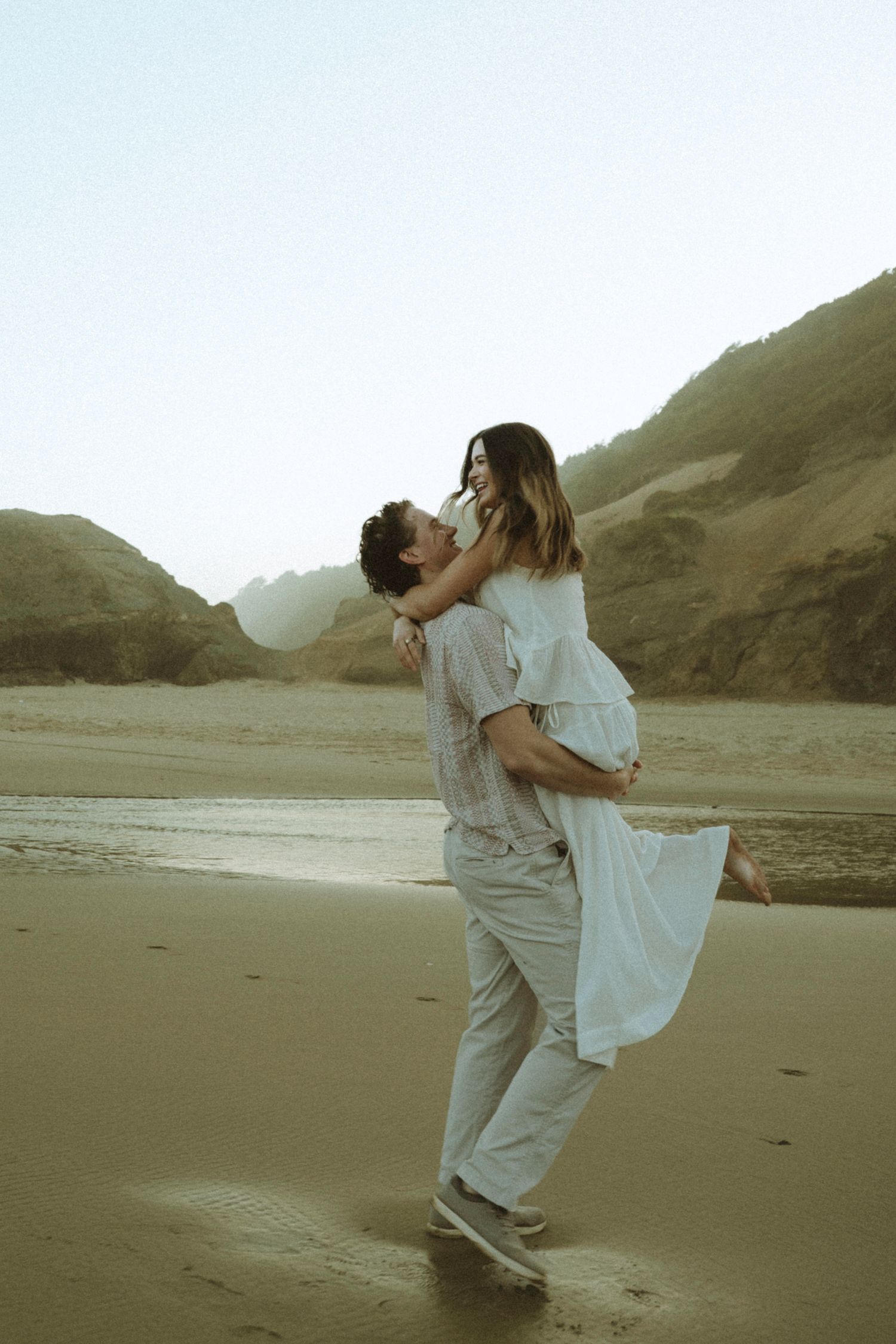 Man lifting woman in white dress on a sandy beach with rocky cliffs in the background.