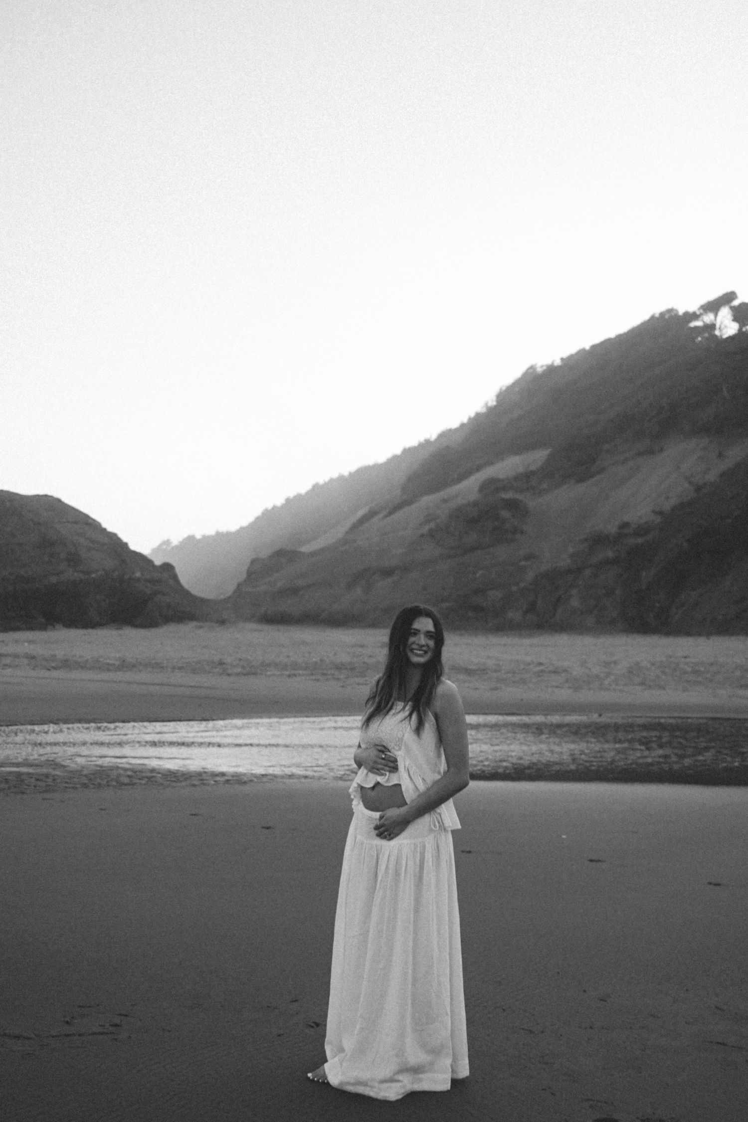Smiling pregnant woman in white maxi dress standing on a beach with mountains in the background, black and white photo.