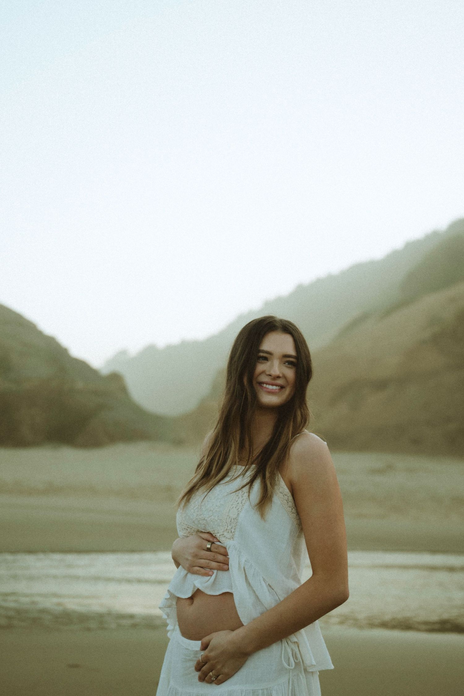 Smiling pregnant woman in white dress posing on a beach with misty mountains behind her.