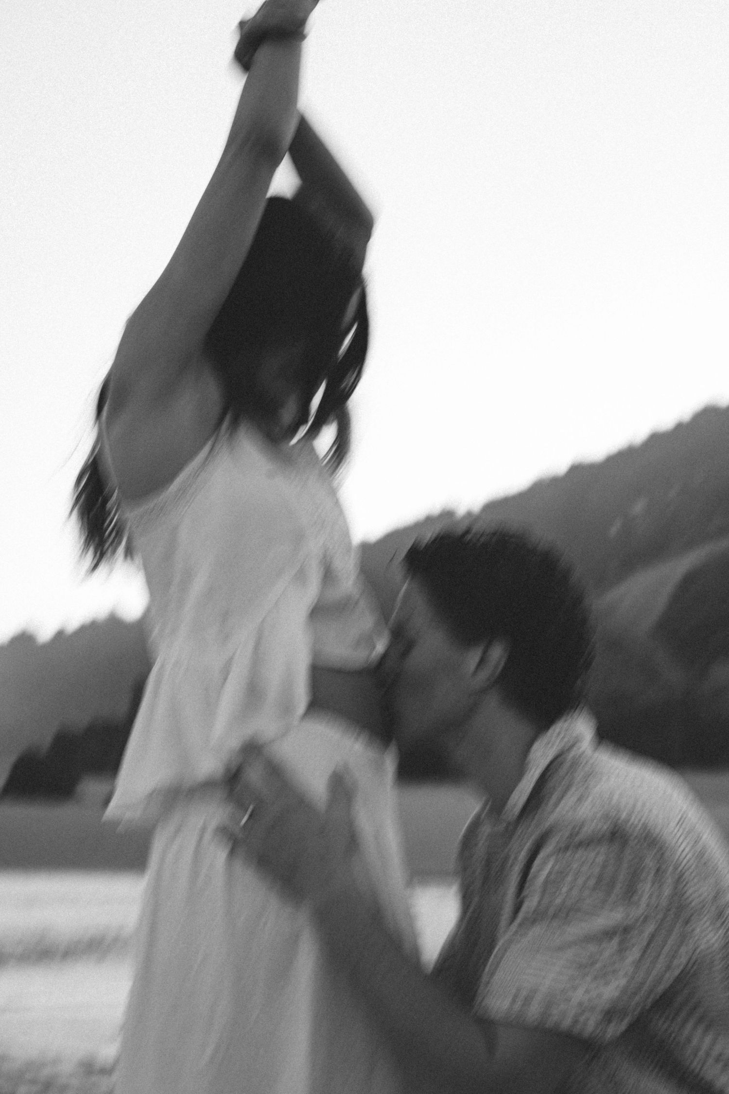 Blurry black and white photo of a man lifting a woman outdoors near a lake with mountains in the background.