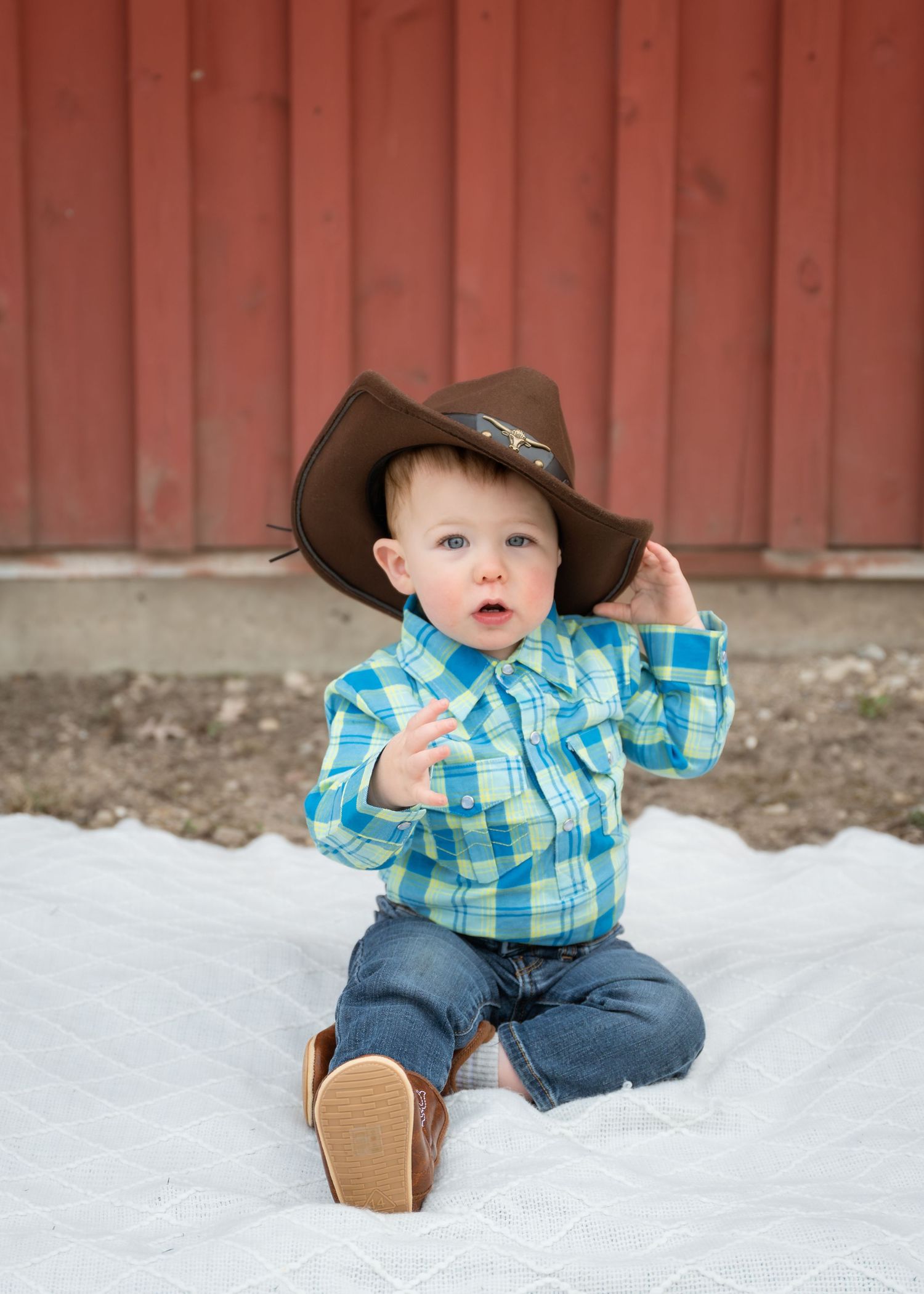 Adorable baby boy in brown cowboy hat, blue plaid shirt, and jeans sitting on white blanket near red barn.