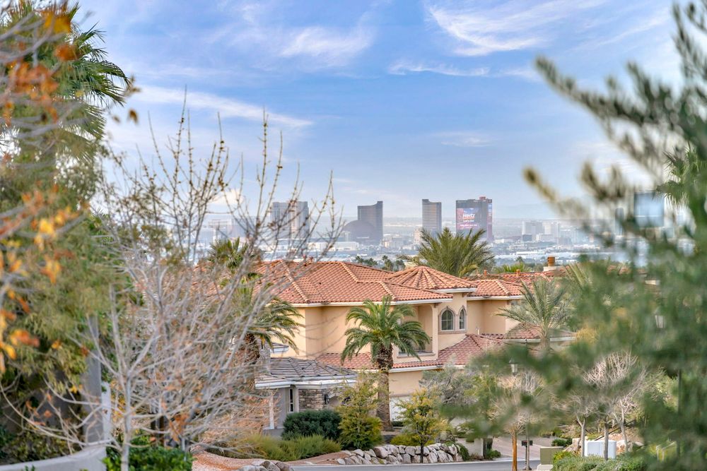 Distant Las Vegas Strip skyline visible through lush trees in an affluent hillside neighborhood.