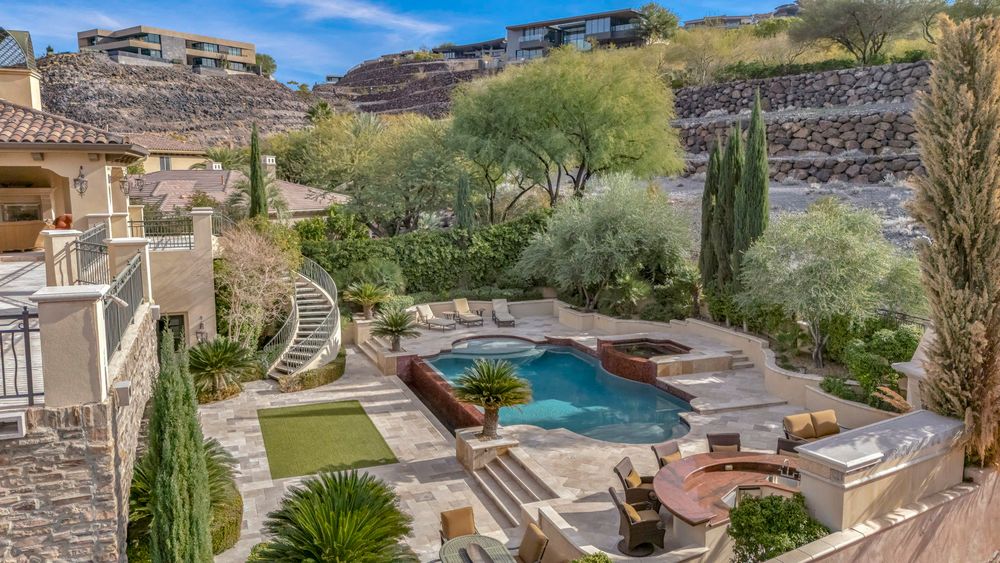 Elegant backyard oasis featuring a pool, spa, putting green, and lush landscaping against rocky hillside.