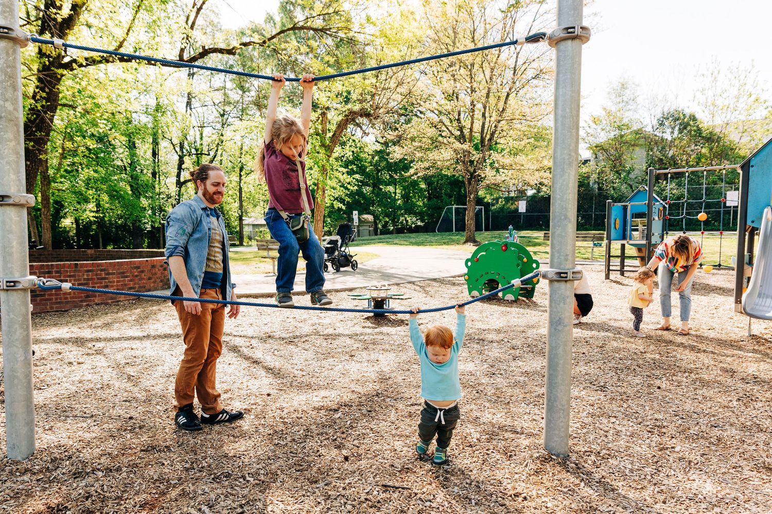Family enjoying a sunny day at a playground with children playing on monkey bars amid green trees.