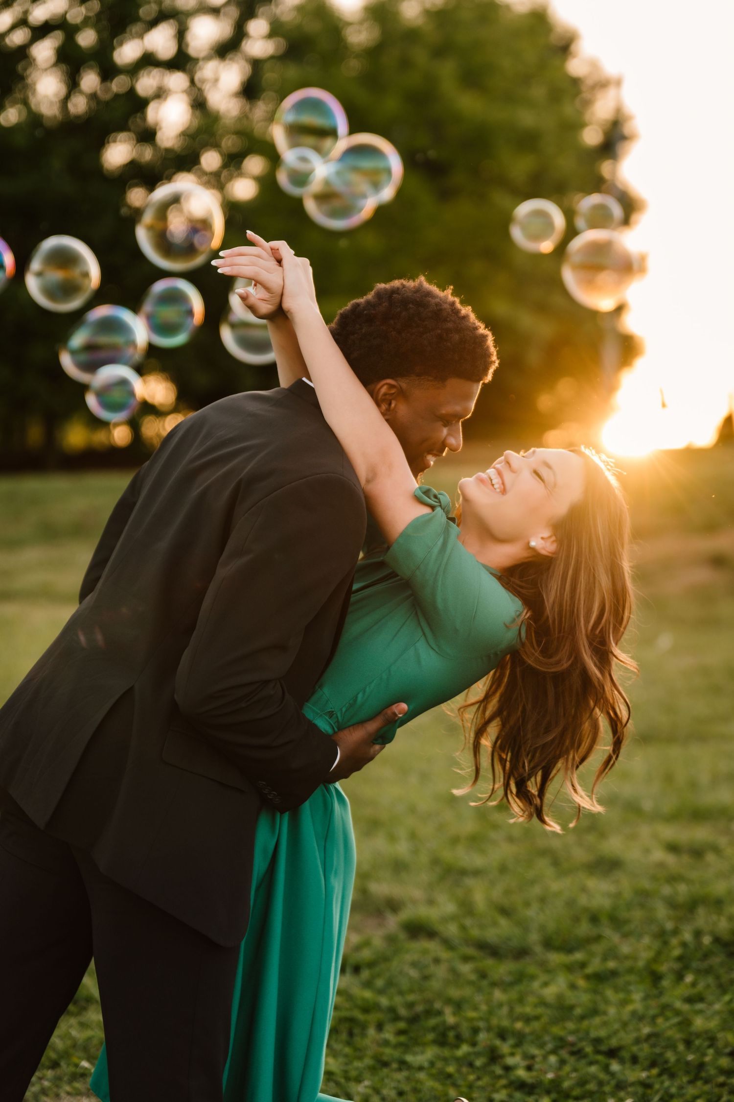 Couple dancing romantically at golden hour sunset in a park, surrounded by floating bubbles, woman in green dress.
