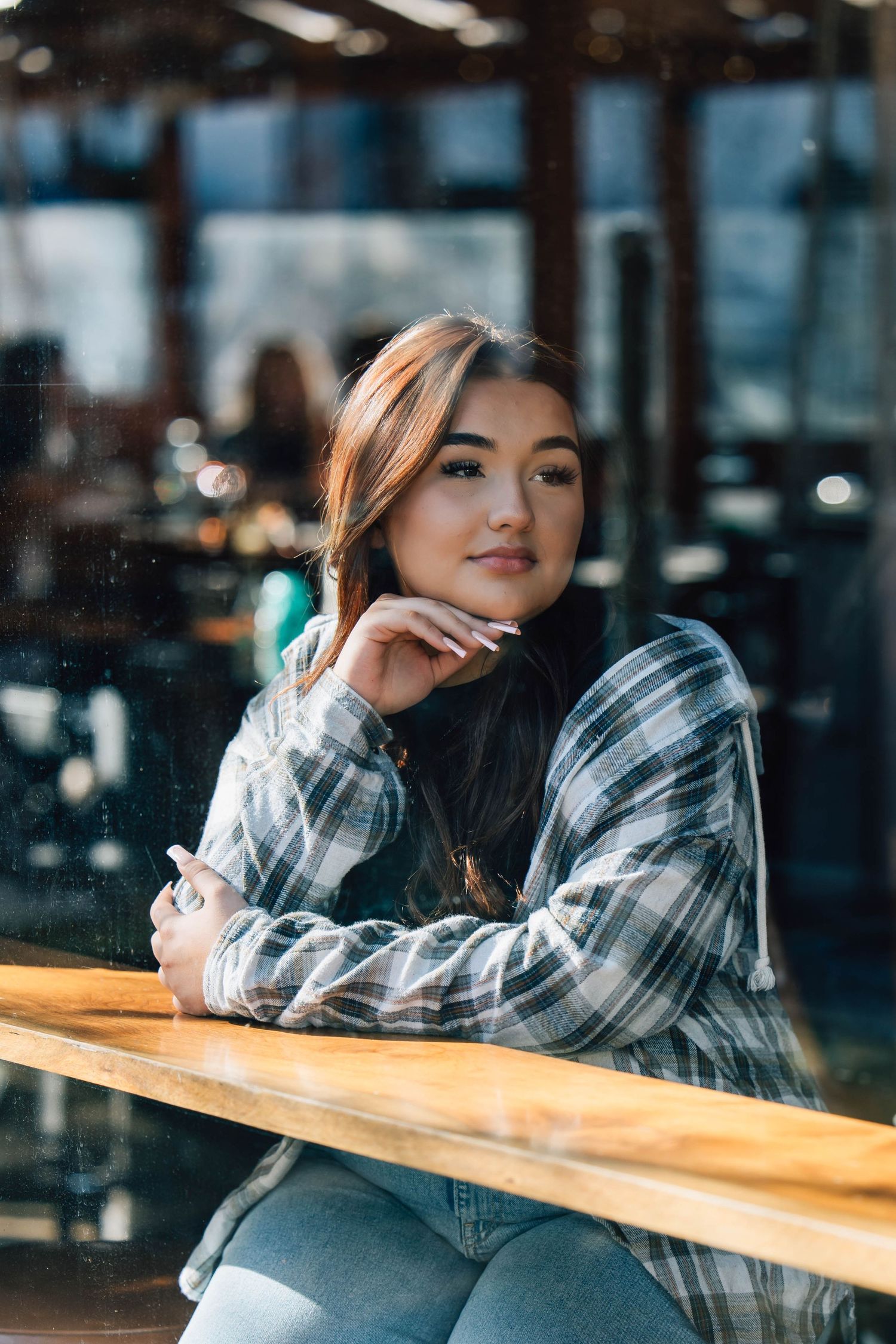 Young woman in plaid shirt sitting at wooden table in cafe viewed through glass window.