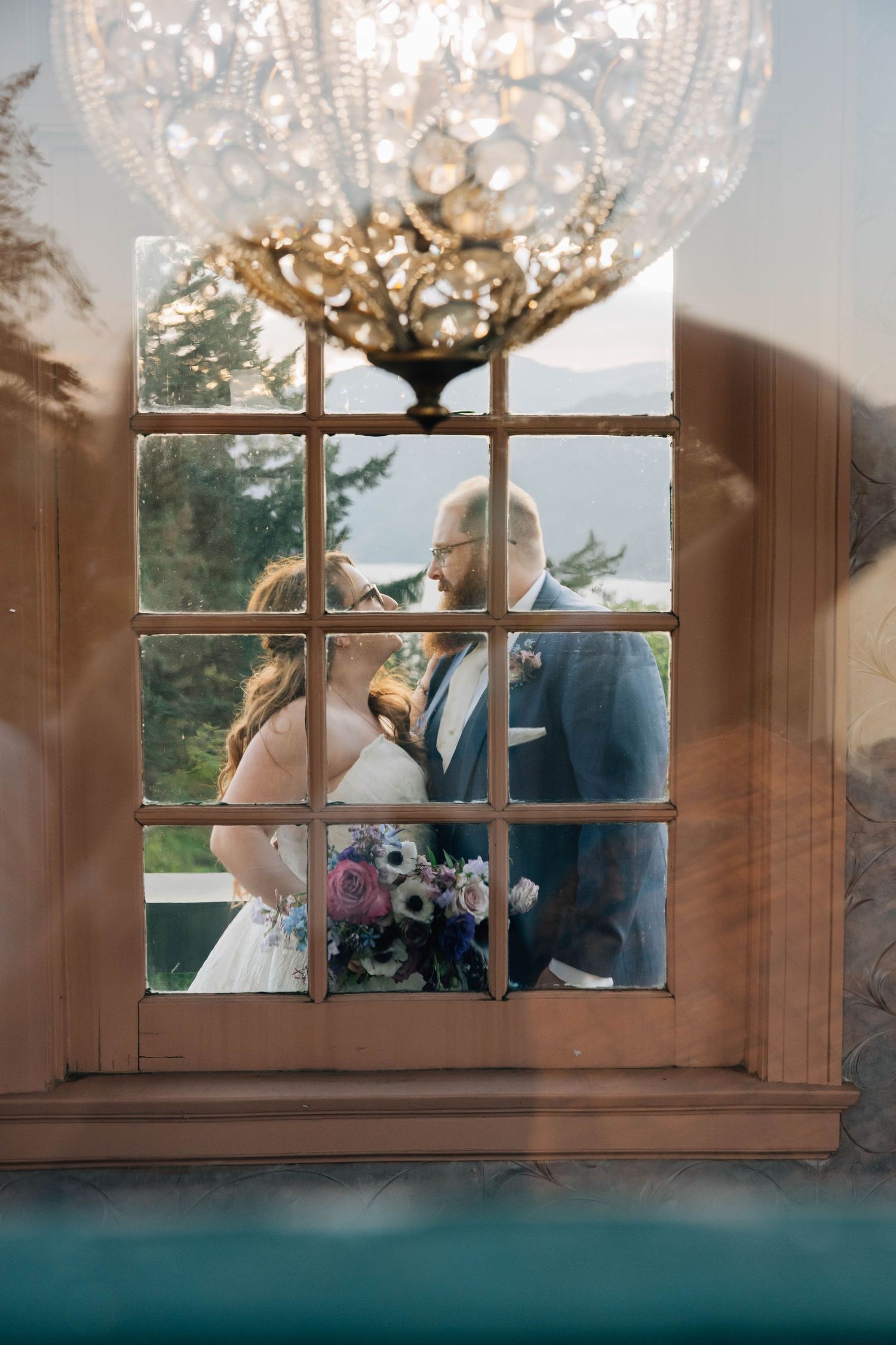Bride and groom sharing a romantic moment viewed through a vintage window with chandelier.