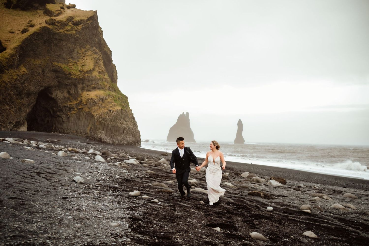 Couple in wedding attire holds hands walking on Iceland black sand beach with dramatic sea stacks and cliffs in background.