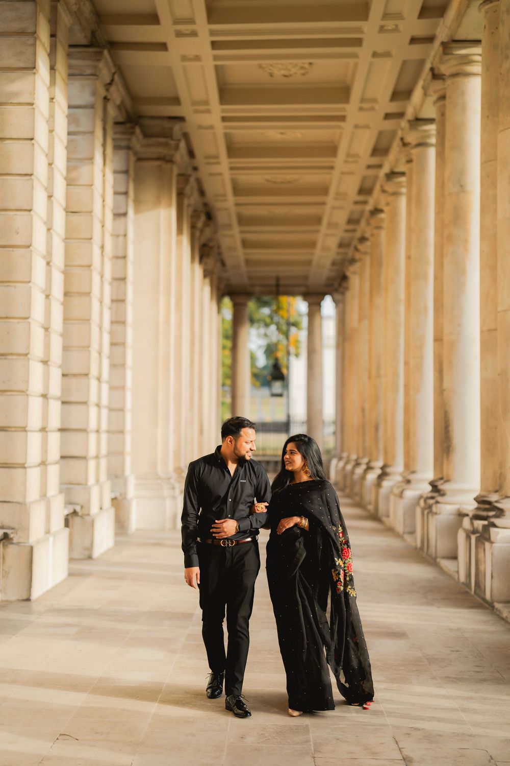 Couple in matching black outfits walking through elegant columned archway, woman in saree, man in suit.