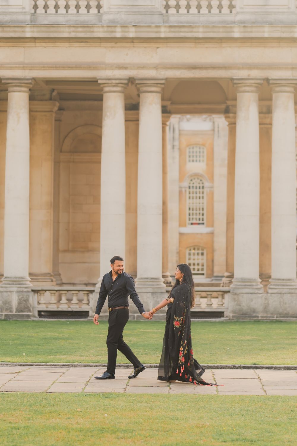 Couple in black outfits holding hands walking in front of grand neoclassical building with tall columns.