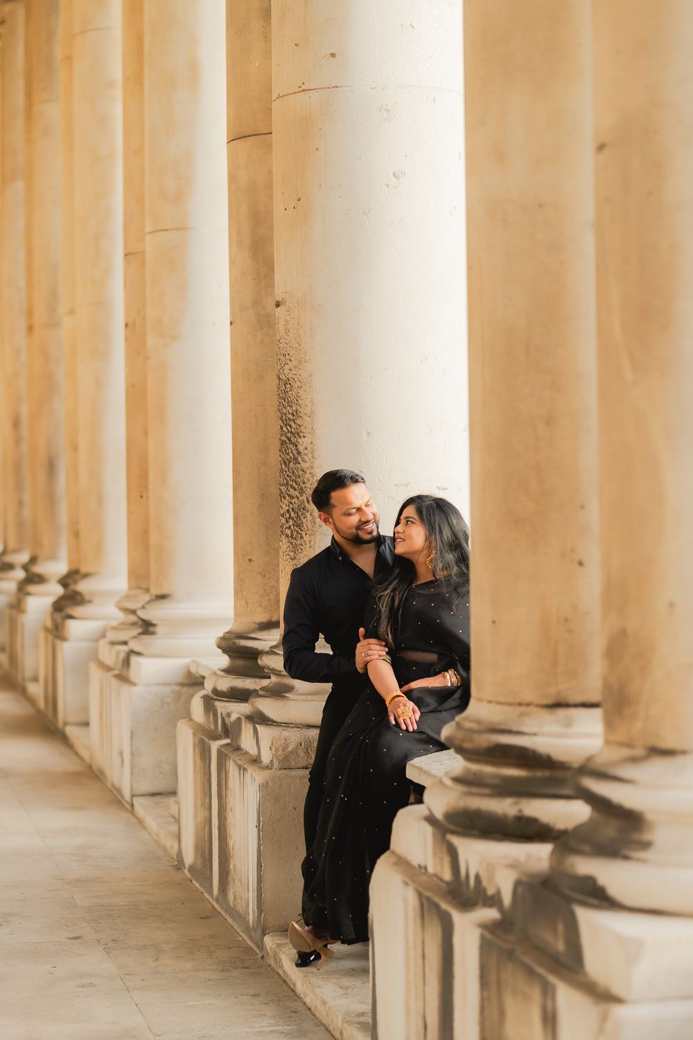 Couple in matching black outfits sharing a romantic moment among grand stone columns of a classical building.