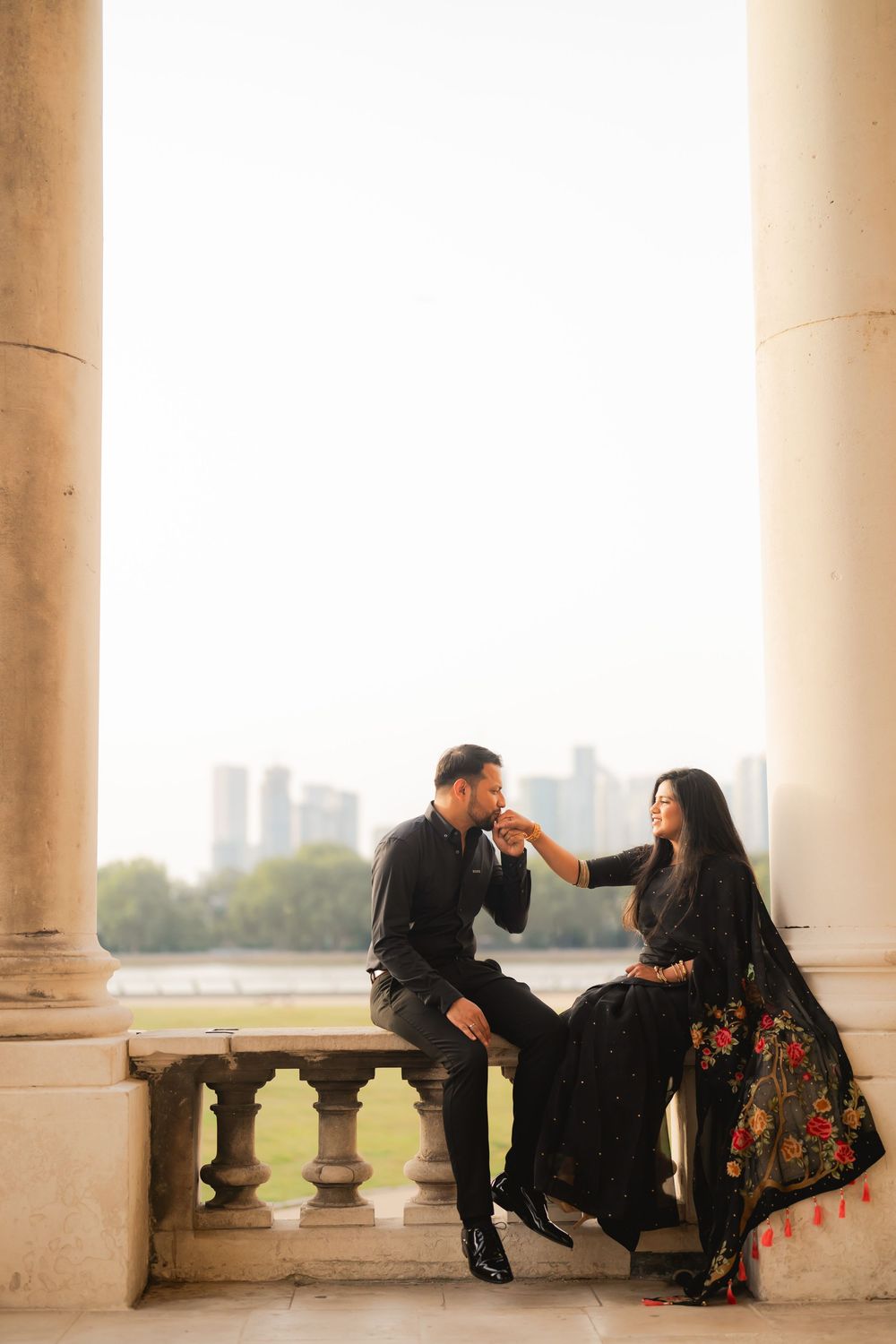 Couple in black attire sharing a romantic moment on a stone balcony with city skyline backdrop.