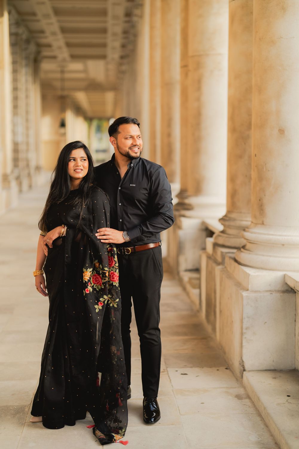 Indian couple in black attire posing elegantly between classical columns in an outdoor colonnade.