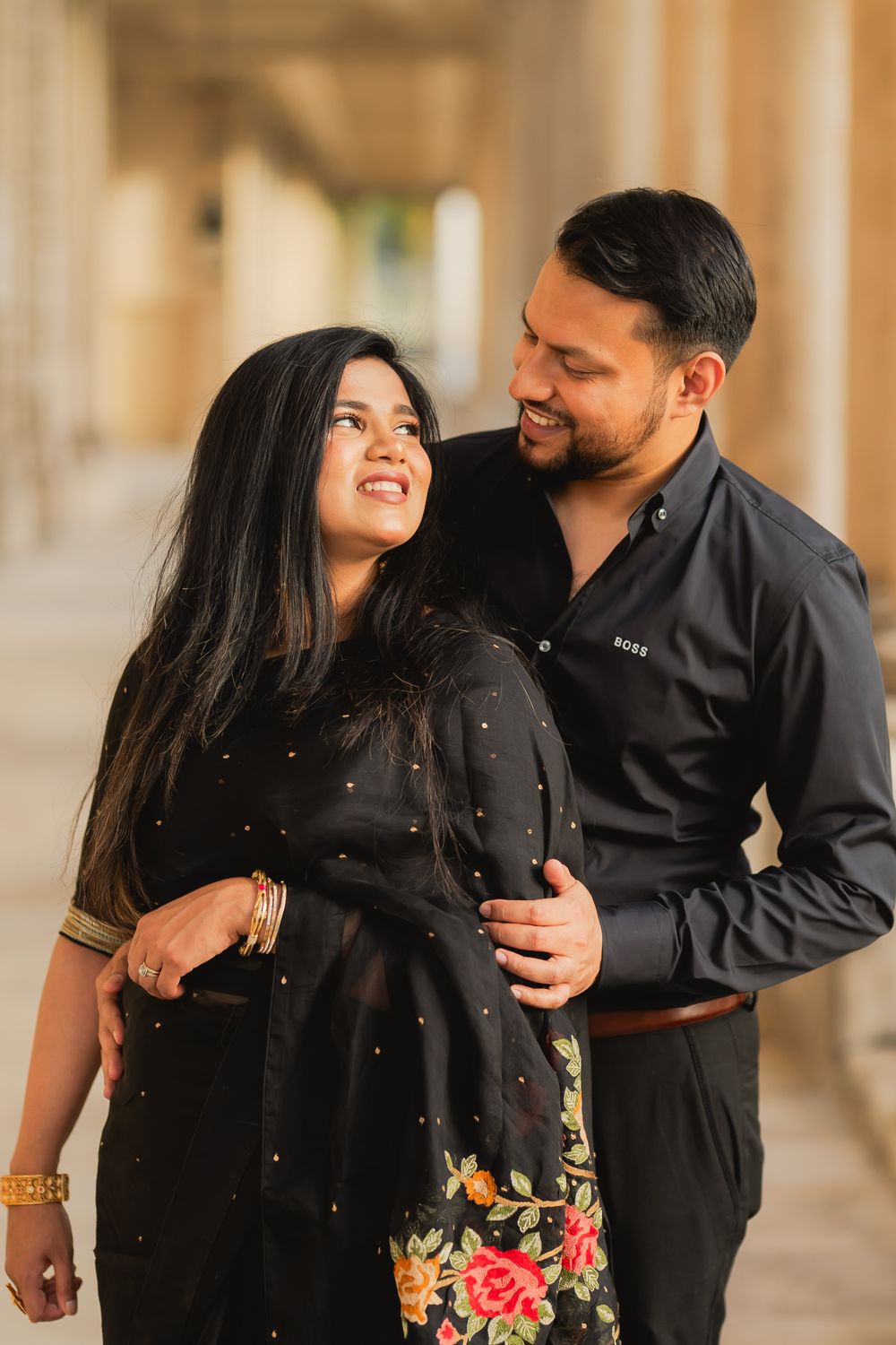 A smiling couple in black outfits share a tender moment, the man embracing the woman from behind in a columned hallway.