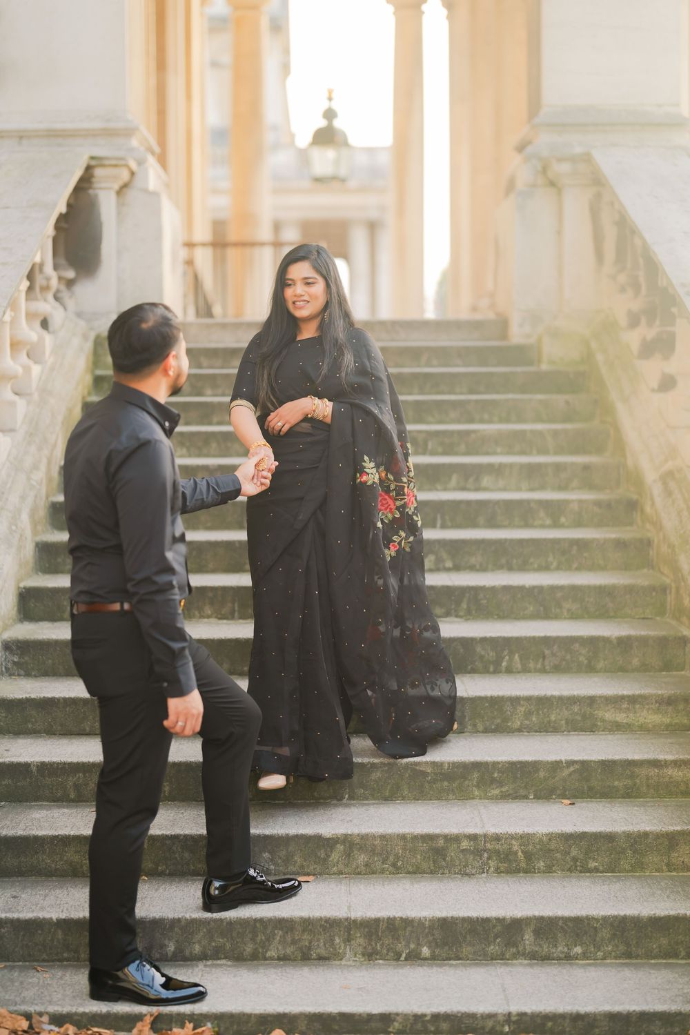 Couple in black attire holding hands on ornate stone steps, woman in embroidered saree, man in formal shirt.