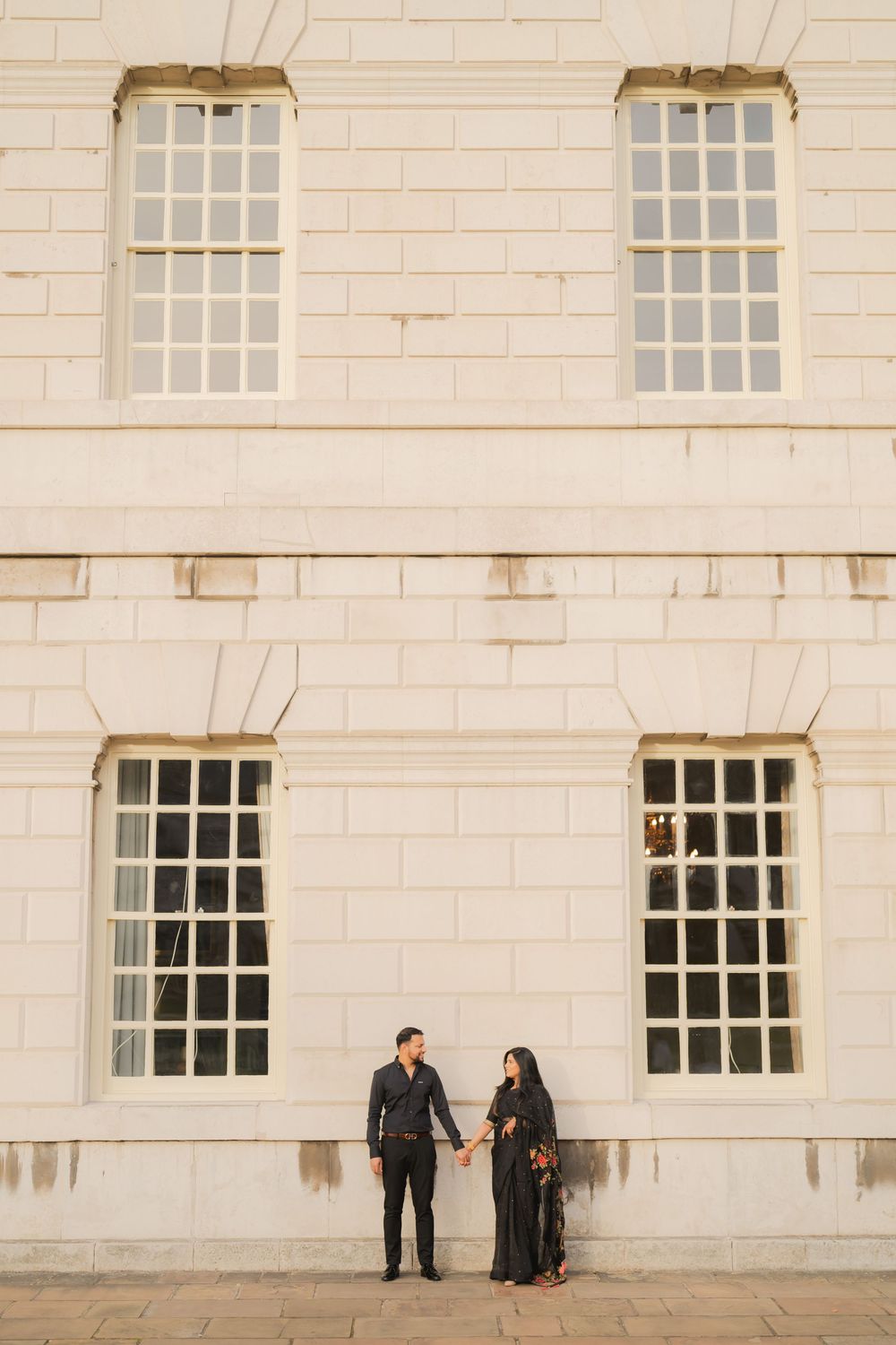Couple in black attire holding hands before a grand white stone building with tall windows.