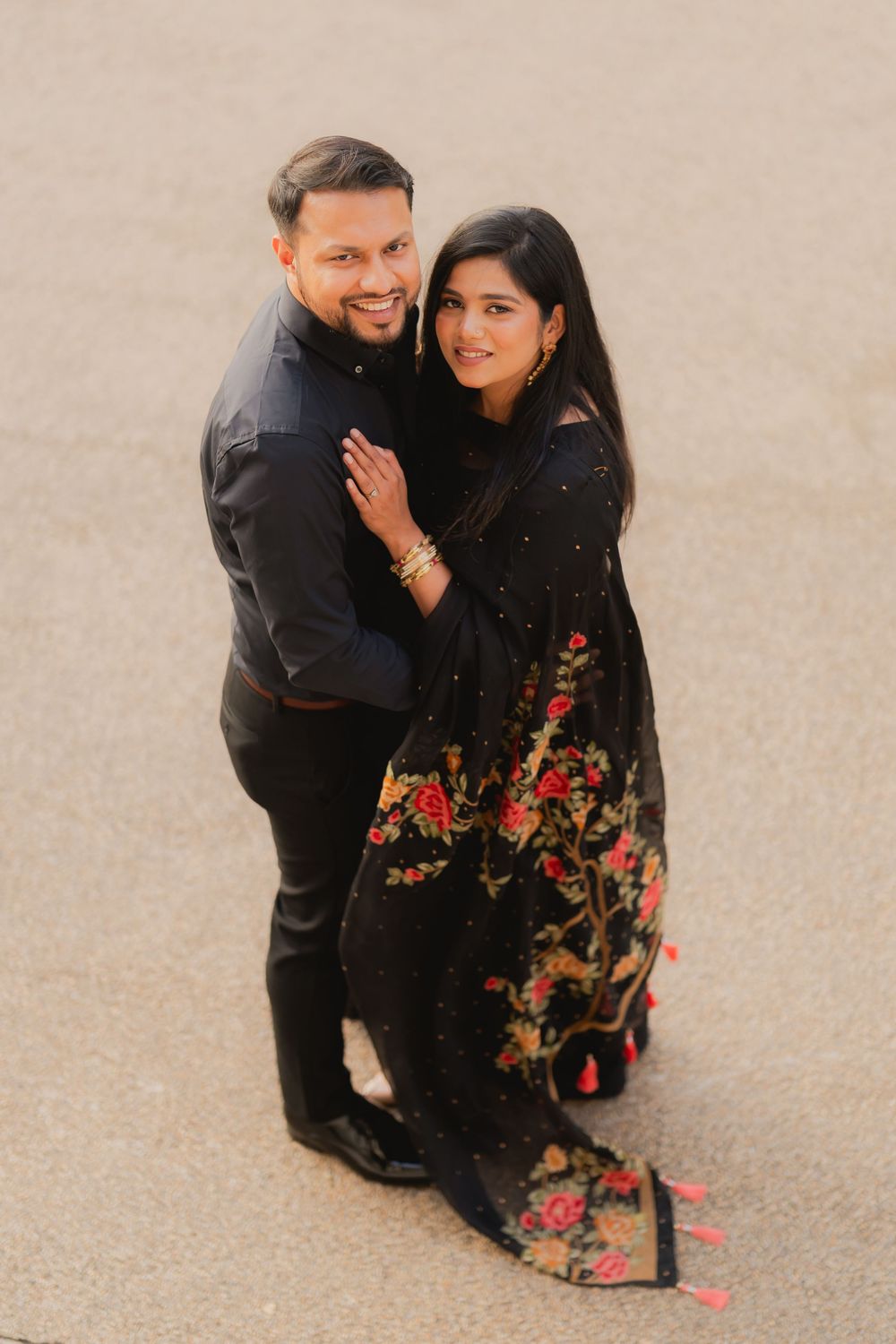 Couple in matching black outfits posing together, woman in floral embroidered anarkali, man in black kurta, smiling at camera.