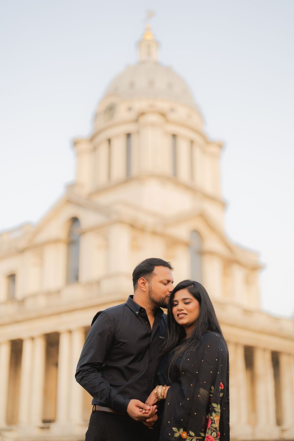 Couple in black attire sharing a romantic moment in front of the Old Royal Naval College dome in Greenwich.