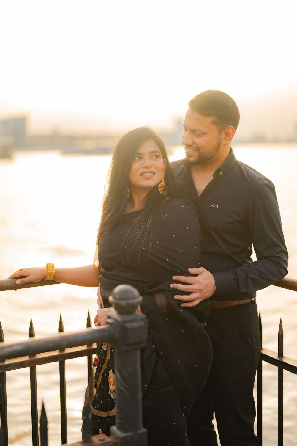 Couple in black outfits embracing by a riverside railing at golden hour sunset.